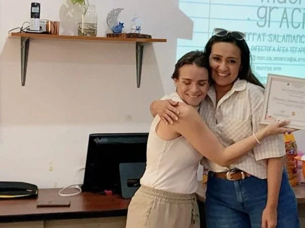 Montse y una Directora de Colegio abrazándose, Montse sostiene un certificado que le fue entregado por su capacitación a docentes.