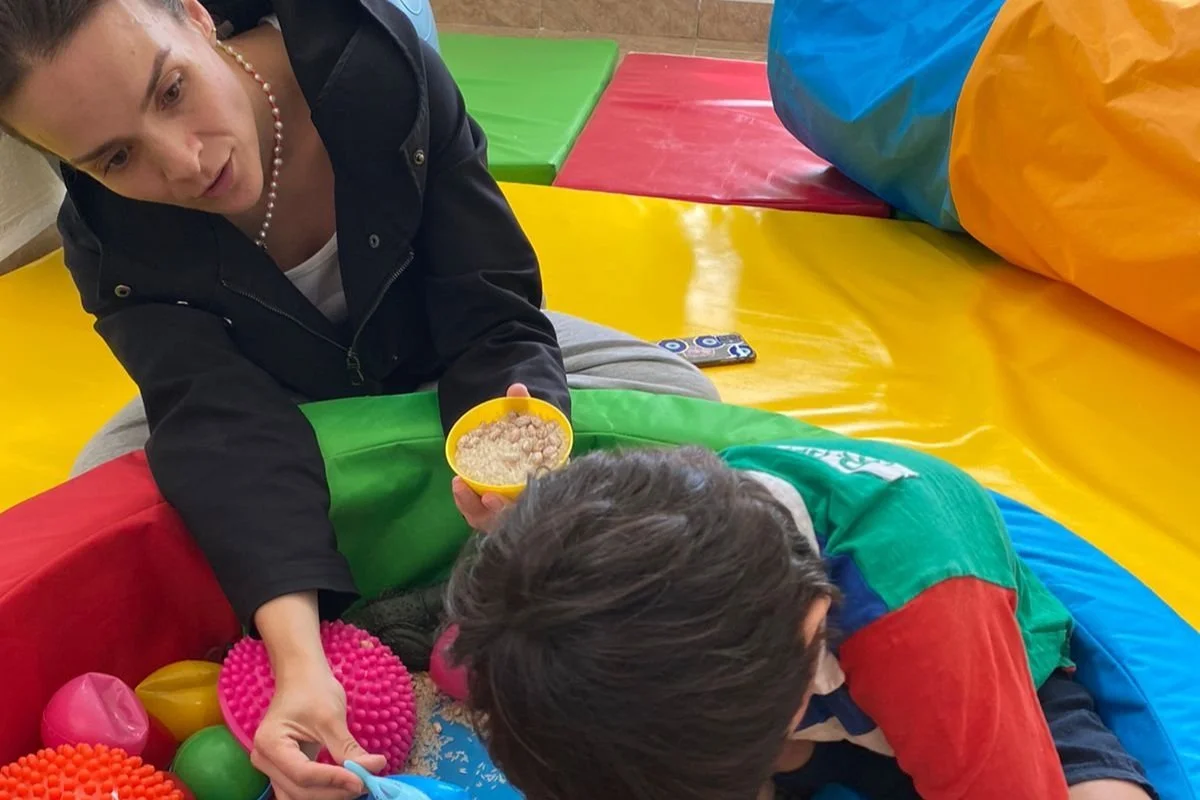 Montse jugando con un niño, donde se encuentran en una alberca rodeada de estímulos sensoriales y colchonetas para apoyar el perfil sensorial del niño.