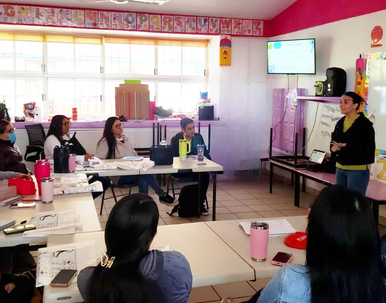 Montse en aula de educación infantil con maestras y directores, pantalla de proyector, hablando sobre la importancia de los apoyos para potenciar la comunicación en el aula.