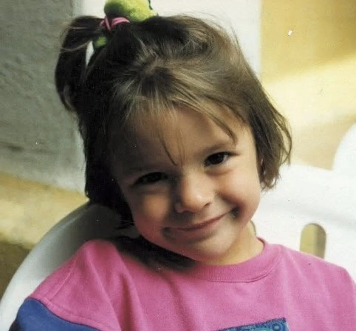 Montse de pequeña, sonriendo, con cabello castaño, lleva un lazo en el cabello y camiseta rosa.