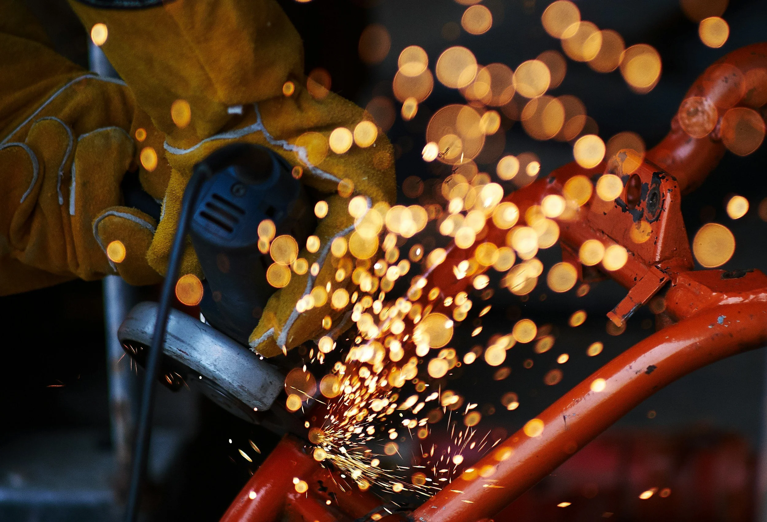 A person wearing yellow gloves uses a grinder on a piece of metal, creating sparks, with a bright bokeh background.