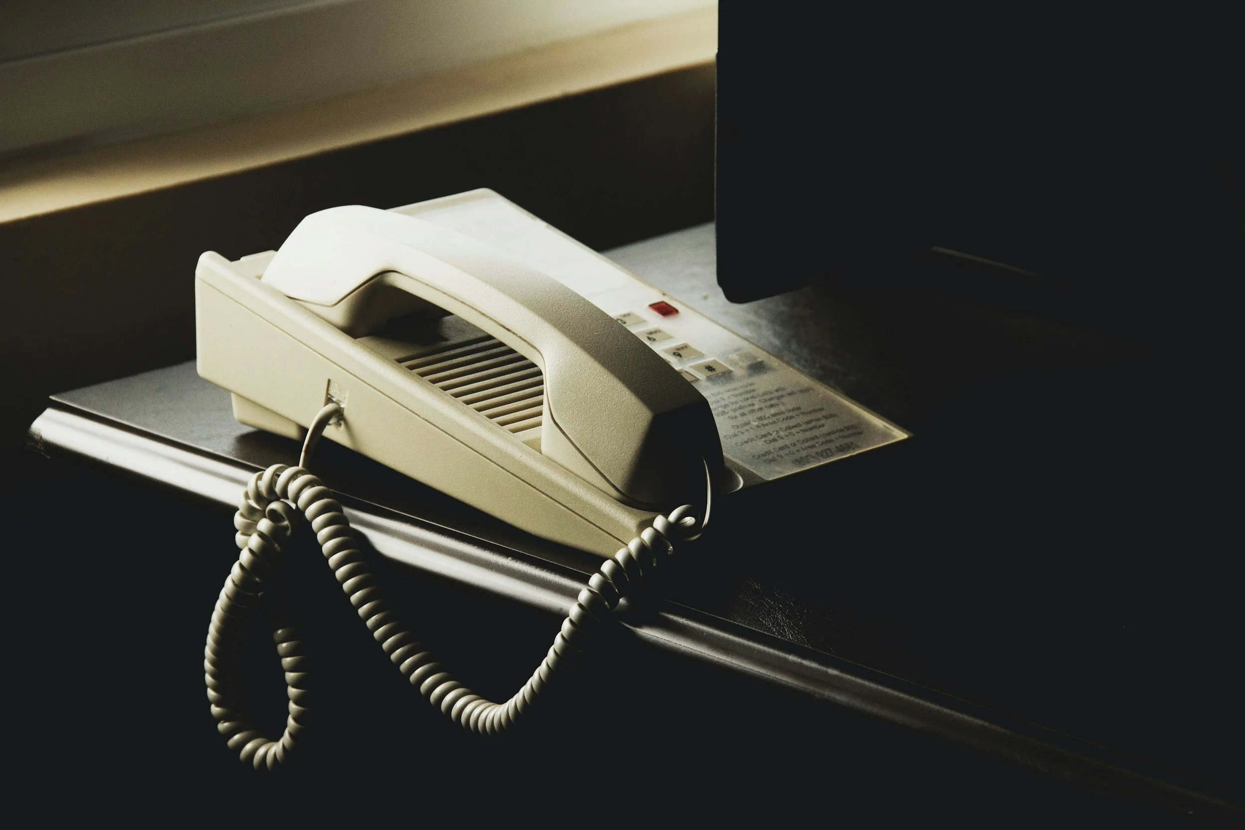 An off-white landline telephone resting on a dark desk near a computer monitor.