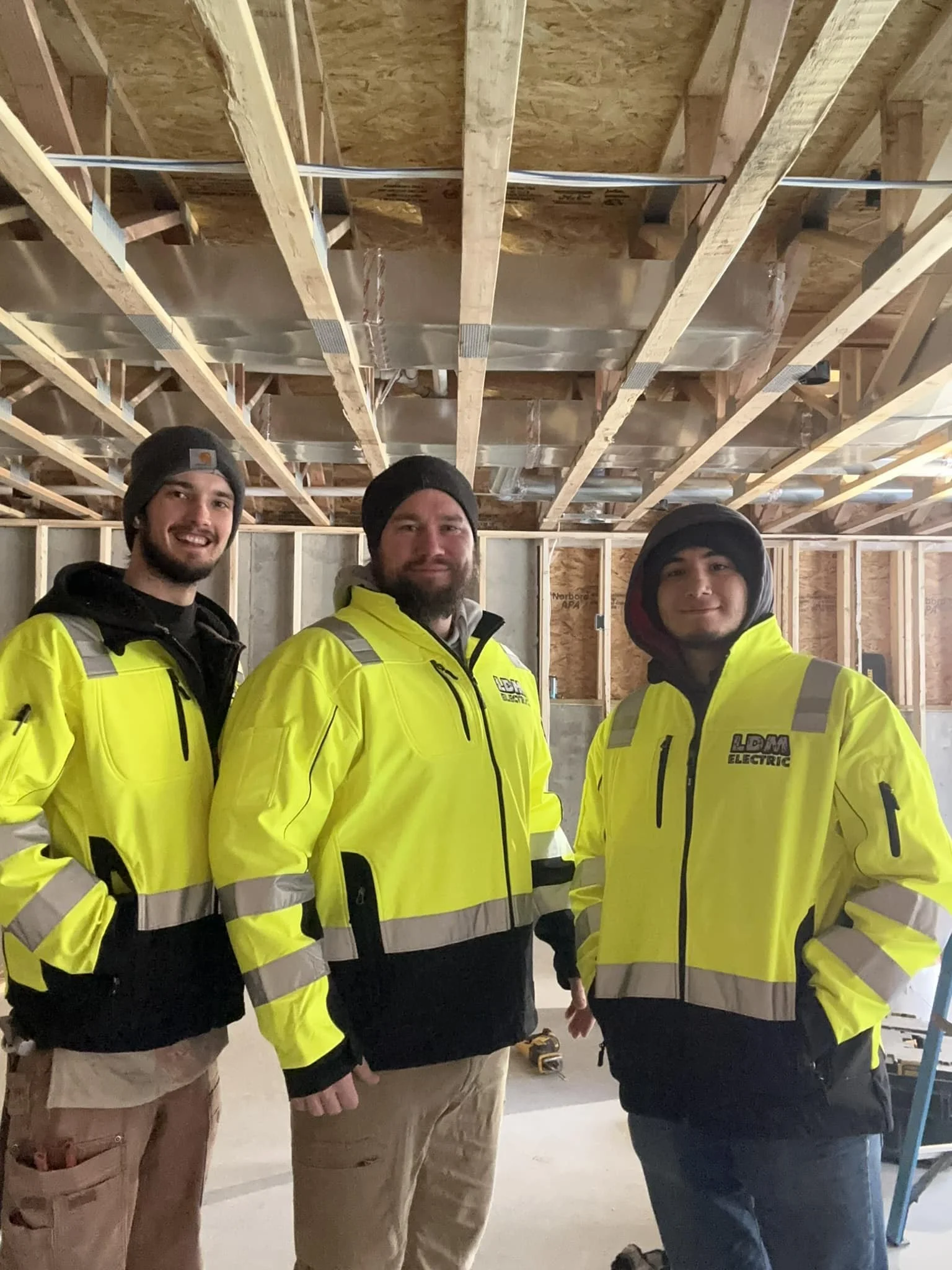 Three construction workers in bright yellow safety jackets posing inside a building under construction, with exposed wooden beams and ceiling framework.