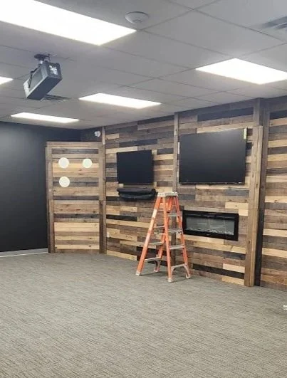 Interior room with wooden accent wall, two large flat-screen TVs, a small shelf beneath one TV, a ladder, and ceiling lights.