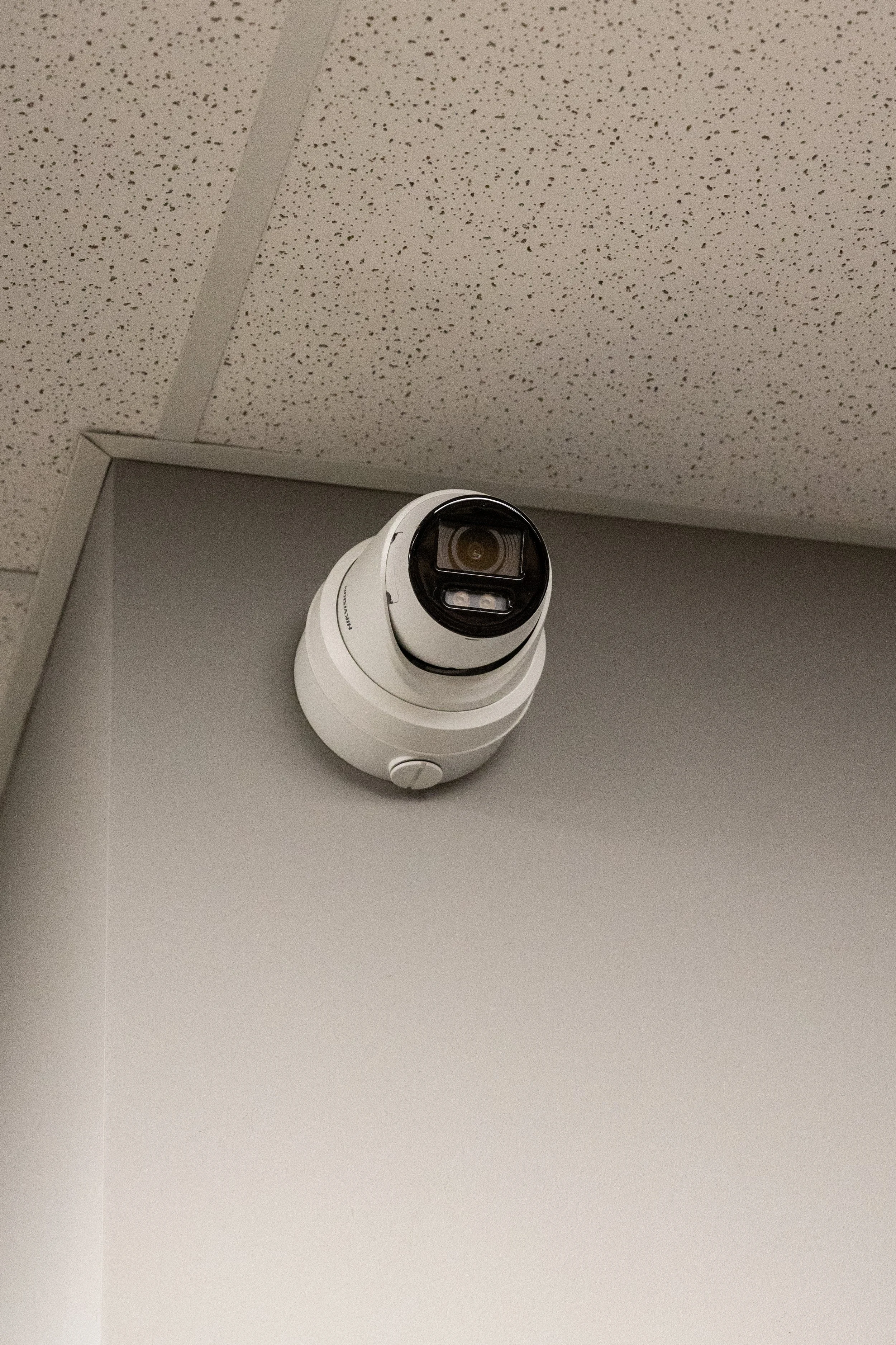 Ceiling-mounted security camera in a corner of a room with acoustic ceiling tiles.