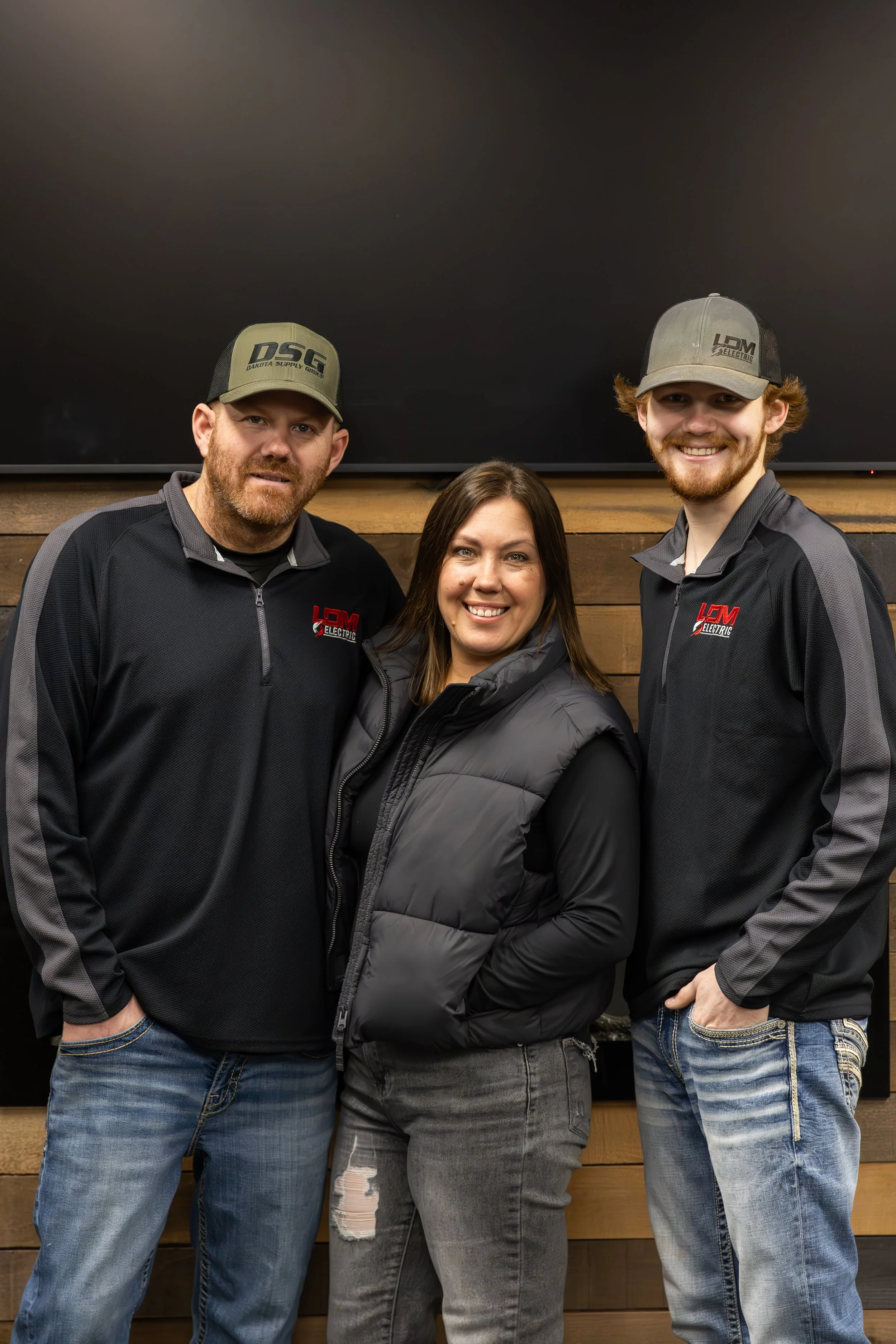 Three people standing together smiling, two men and one woman, wearing casual jackets and caps, in front of a wooden wall with a large black screen.