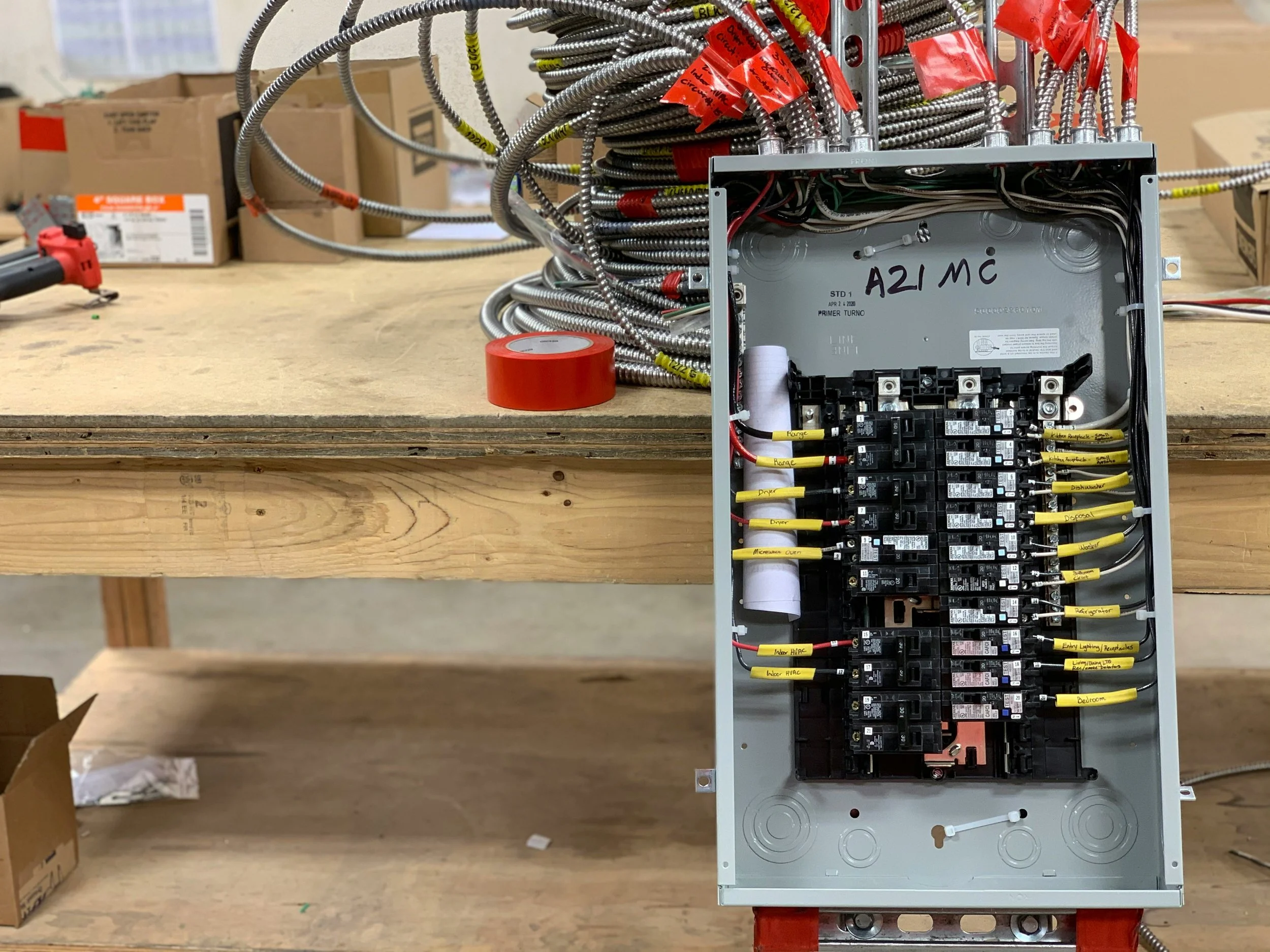 Open electrical circuit box with labeled wires and circuit breakers, resting on a wooden workbench with tools and boxes in the background.
