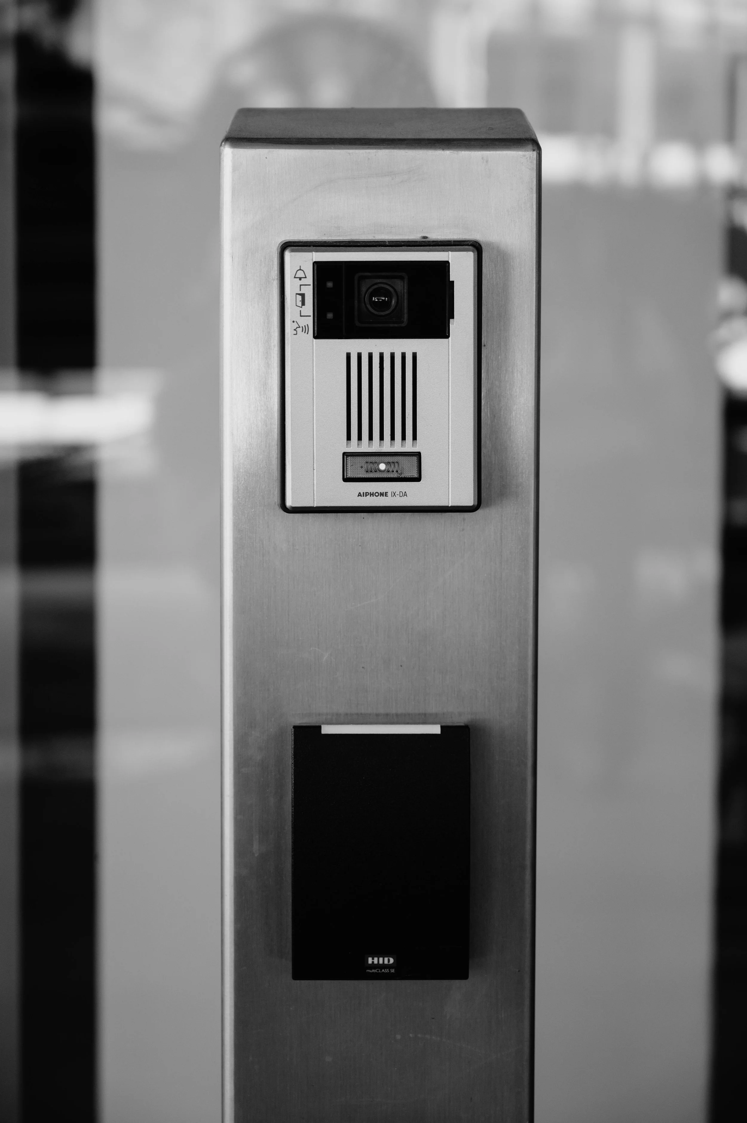 Black and white photo of a metallic security or access control device with a camera and speaker, mounted on a metal post.