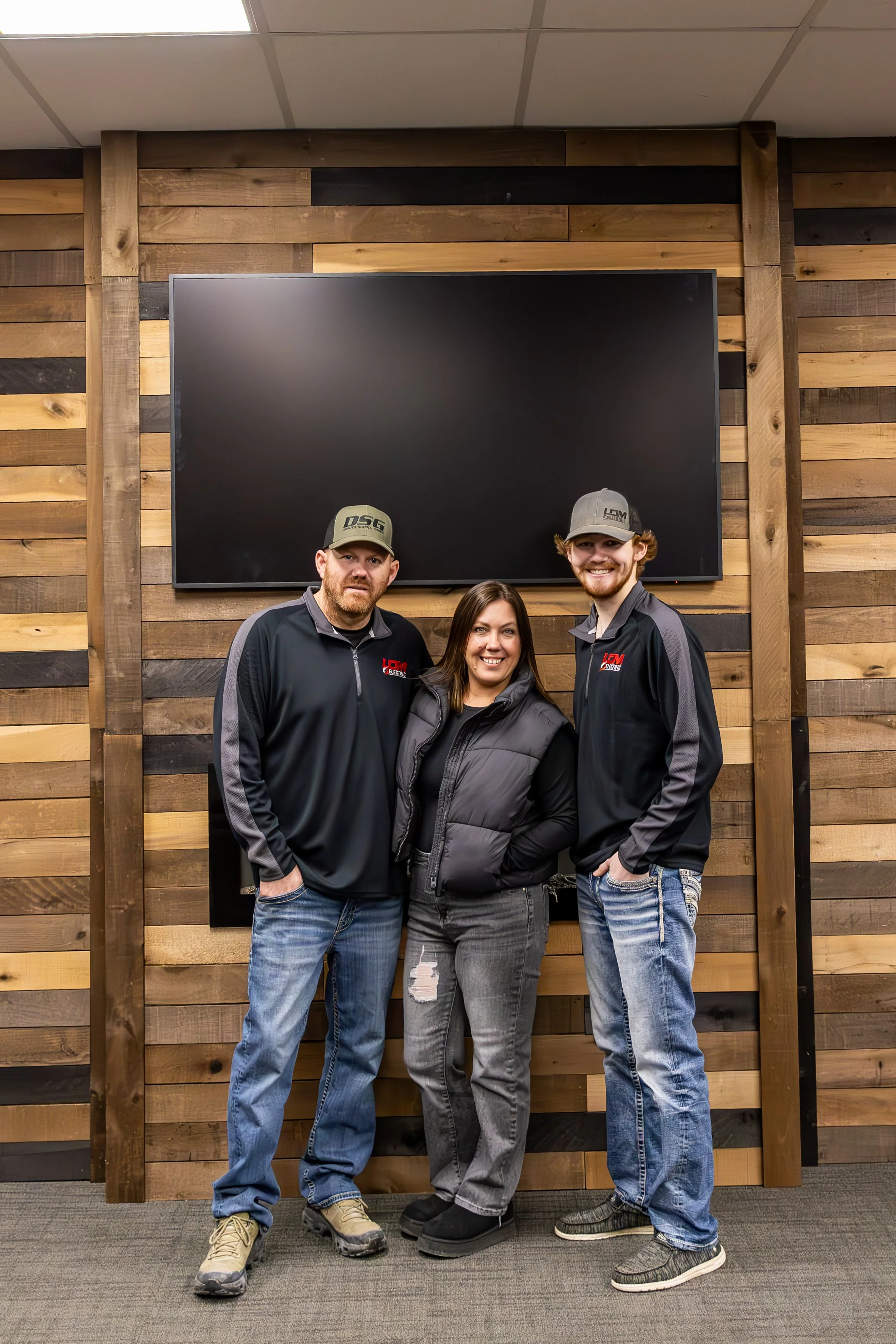 Three people standing in front of a wooden wall with a large black screen.