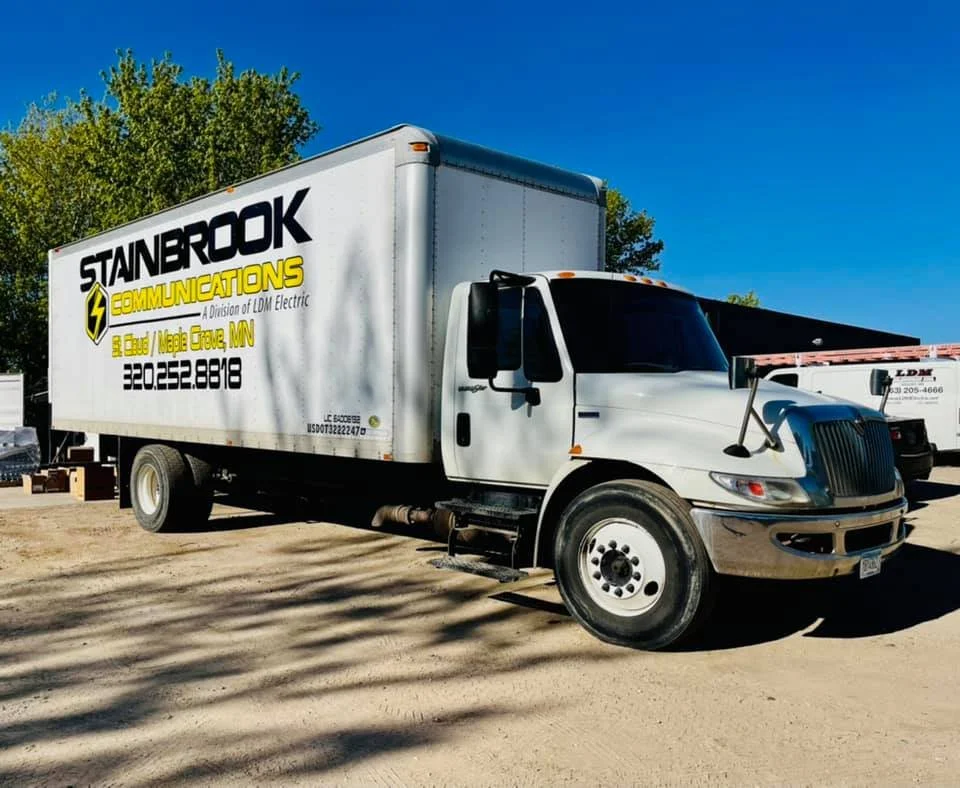 White box truck with advertising for Stanbrook Communications, a division of LDM Electric, parked on dirt ground with trees and other trucks in the background, under a clear blue sky.