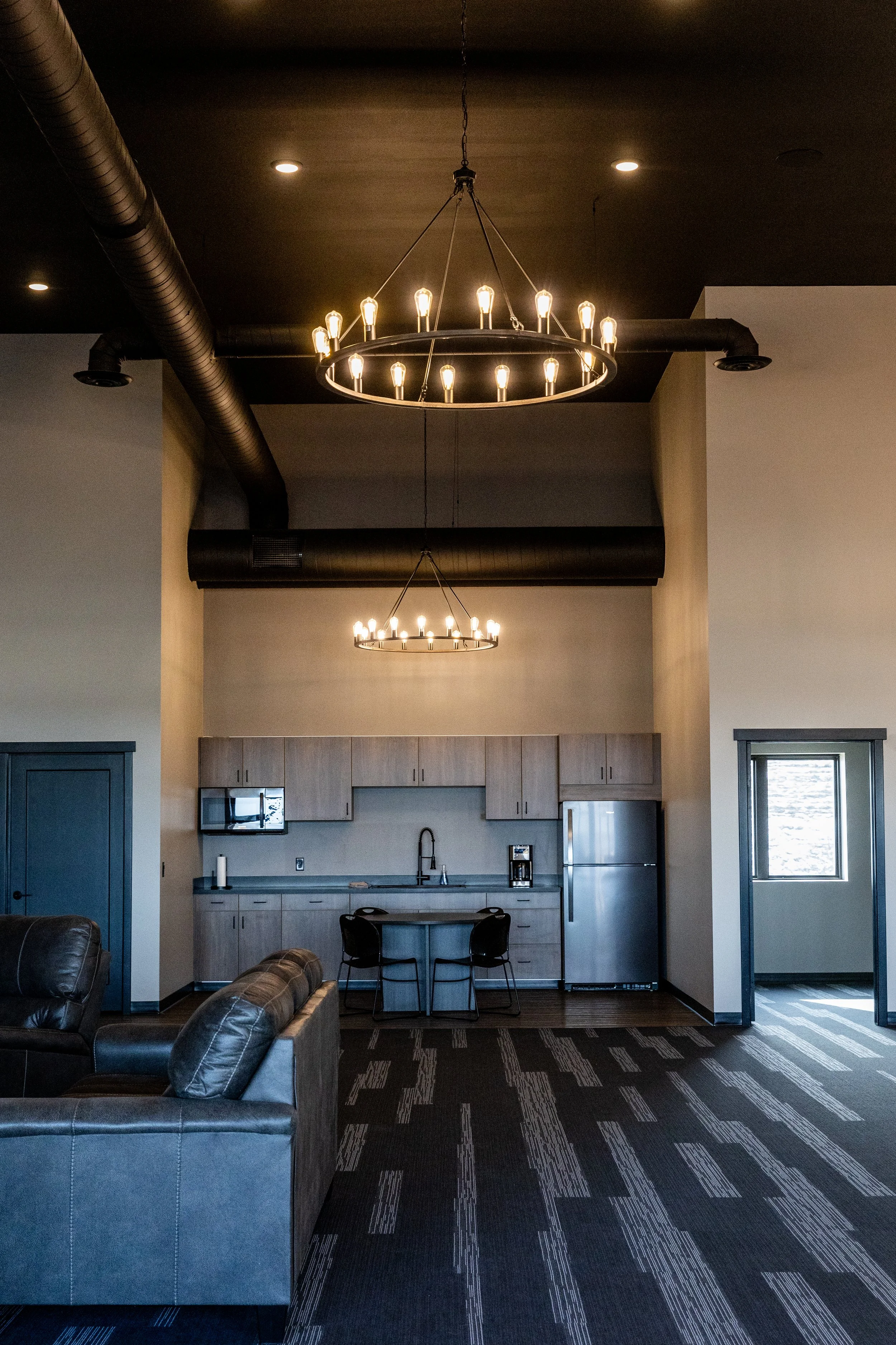 Interior of an apartment or lounge area with high ceilings, black lighting fixtures, a kitchenette with a refrigerator, microwave, and sink, a small table and two chairs, a leather sofa, a window, and dark patterned carpet.