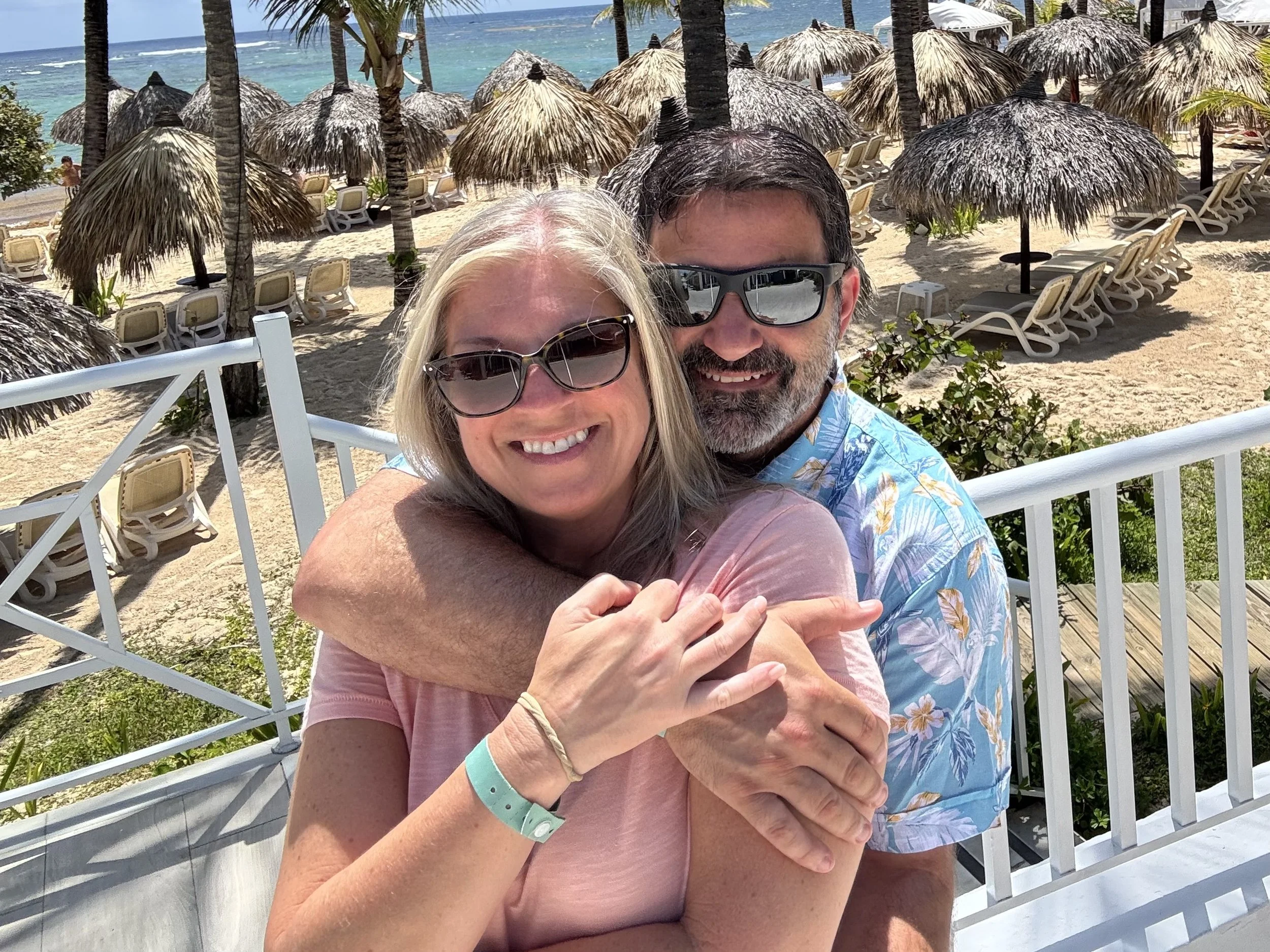 A smiling couple hugging, outside at a beach resort with thatched umbrellas and lounge chairs, palm trees, and an ocean in the background.