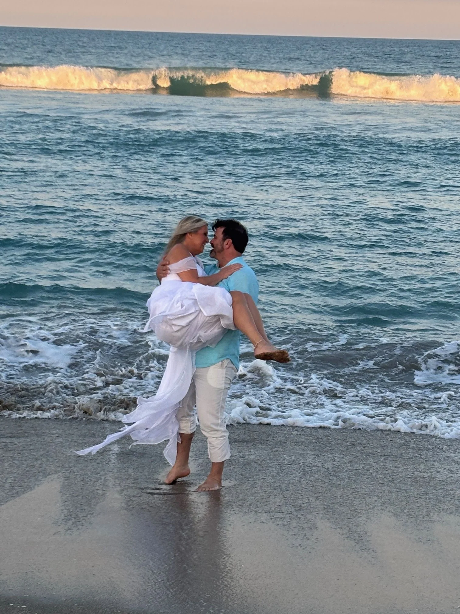 A couple is on the beach, with the man lifting the woman in his arms as they share an intimate moment, with the ocean waves and a sunset in the background.