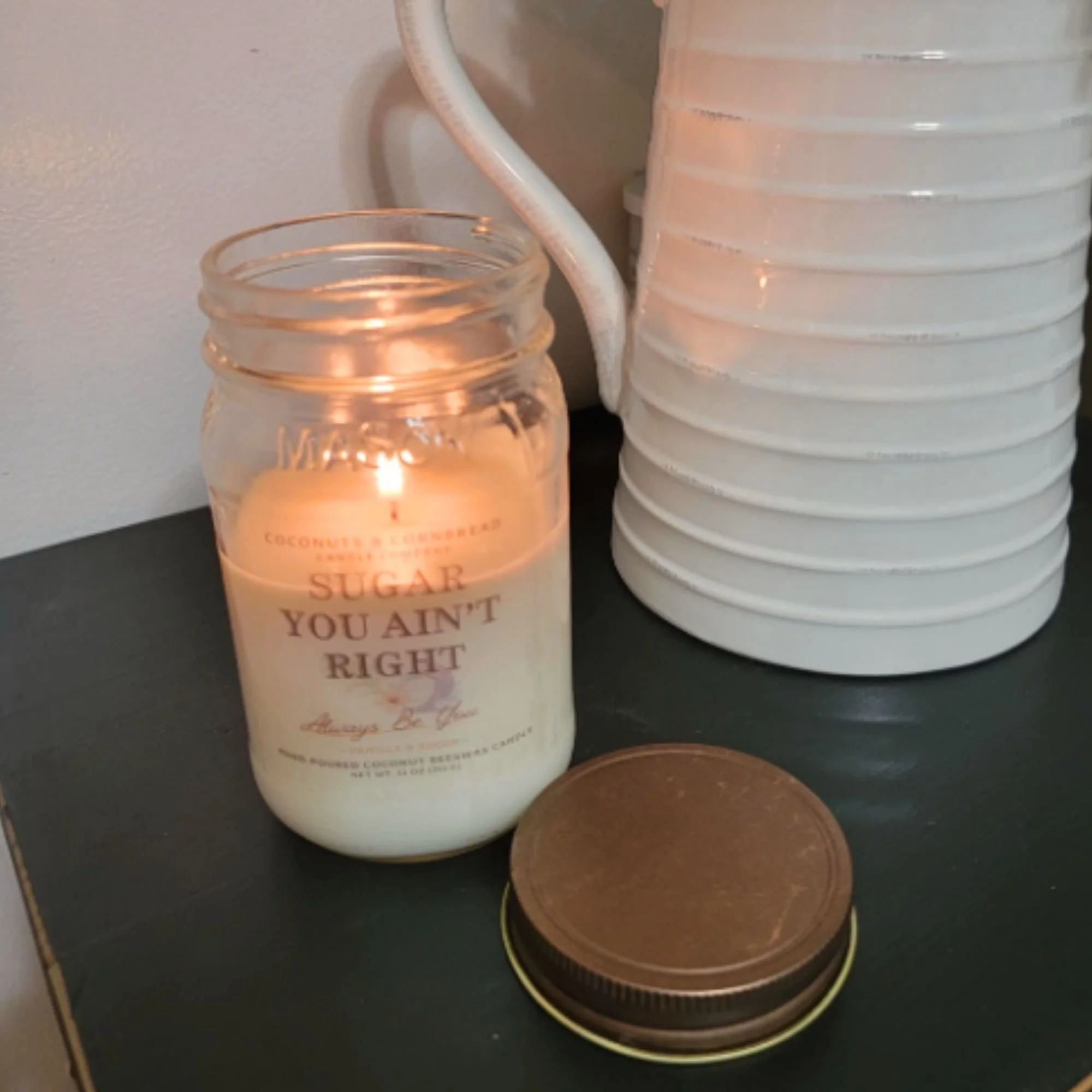 Mason jar with a lit candle inside, next to a white vase of flowers, and a mason jar lid, on a black surface against a plain wall. The candle is called Sugar You Ain't Right, a Vanilla candle by Coconuts and Cornbread Candle Company.
