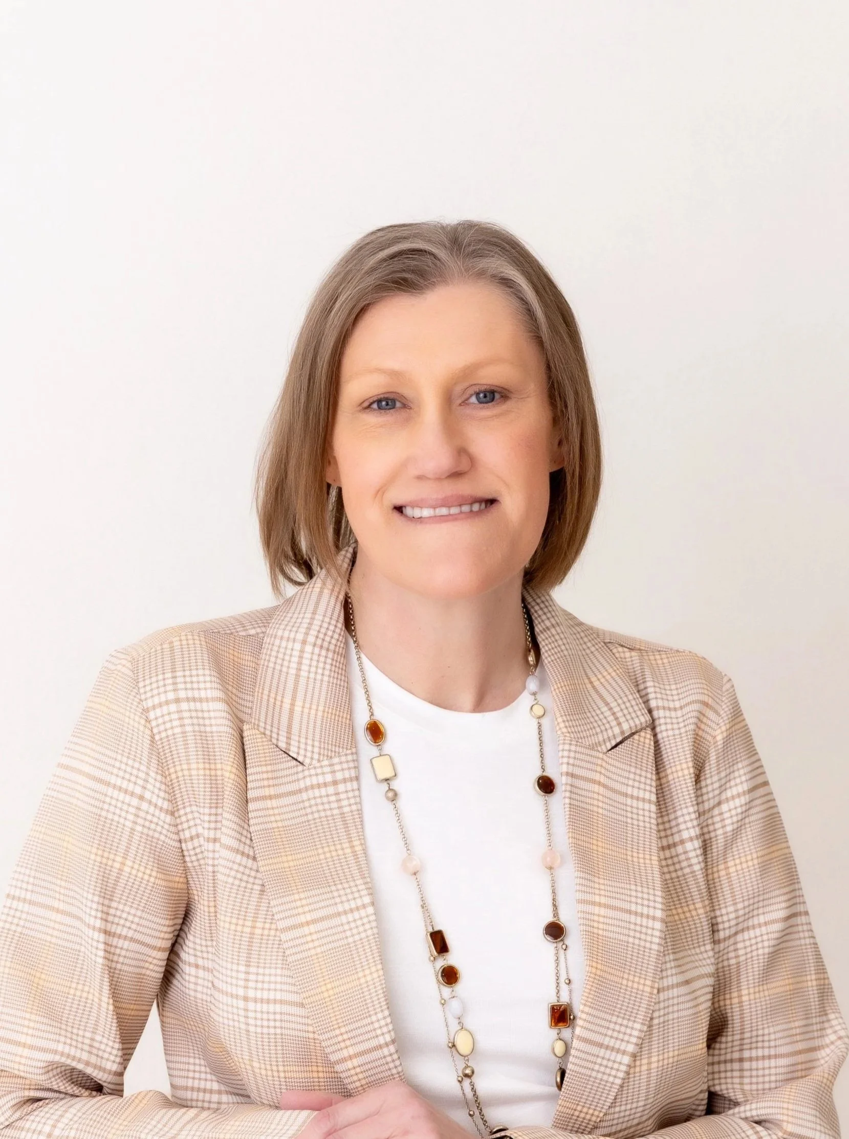 Dessie Calihou with shoulder-length light brown hair, wearing a beige checkered blazer over a white top and a long necklace with various pendant shapes, standing against a plain white background.