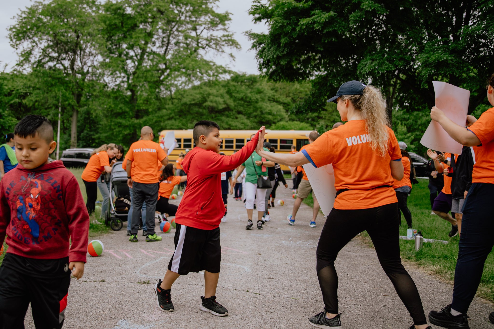 A woman volunteer high-fives a young boy at an outdoor community event with many children and adults participating, with trees and a yellow school bus in the background.