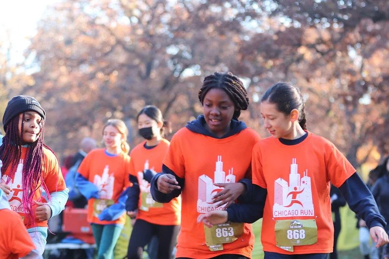 Group of diverse women running together in a park during a Chicago Run event, all wearing orange shirts with a runner logo and bib numbers.