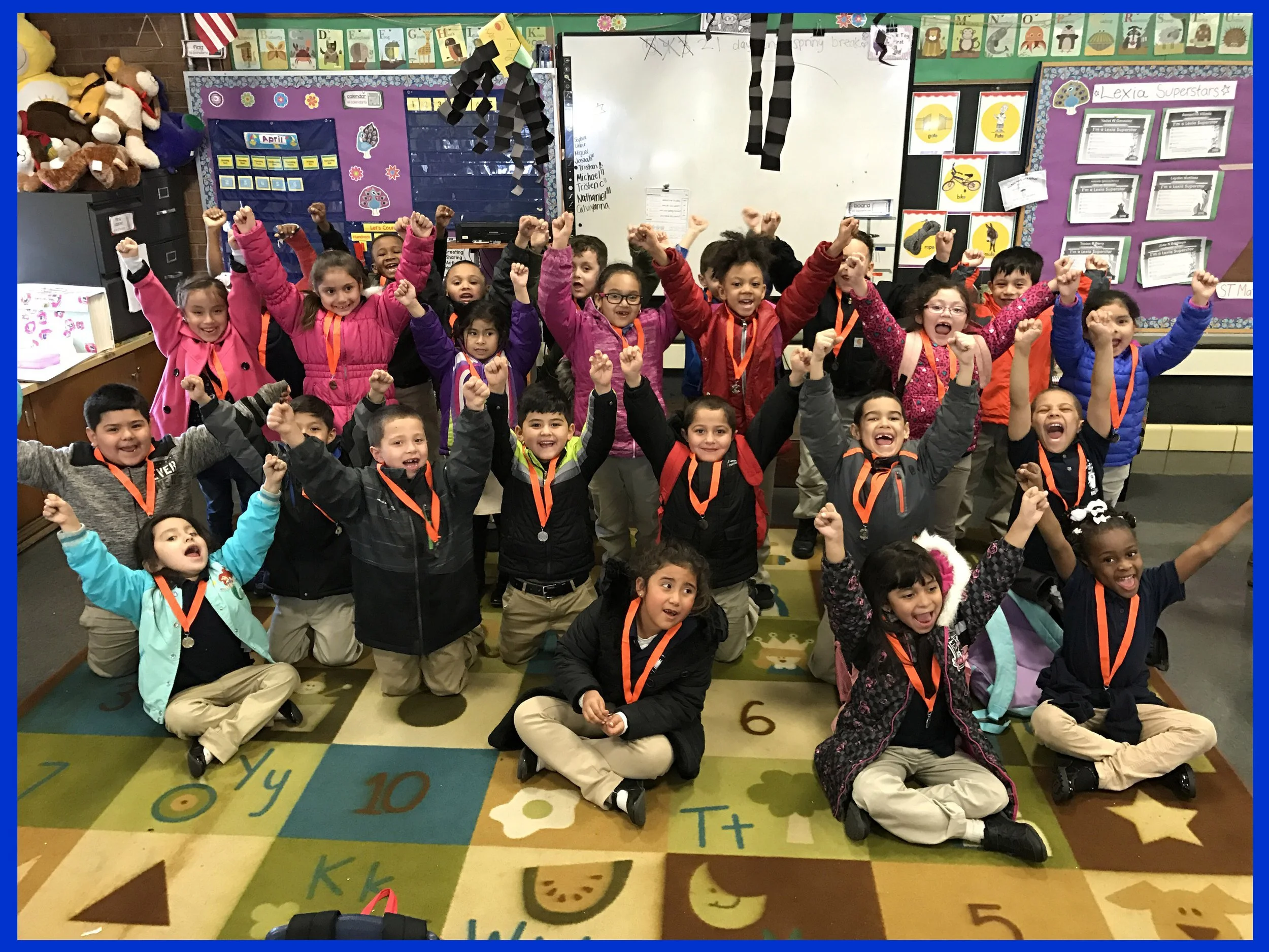 A group of young students in a classroom celebrating with raised arms and joyful expressions, wearing medals around their necks, on a colorful alphabet and number rug.