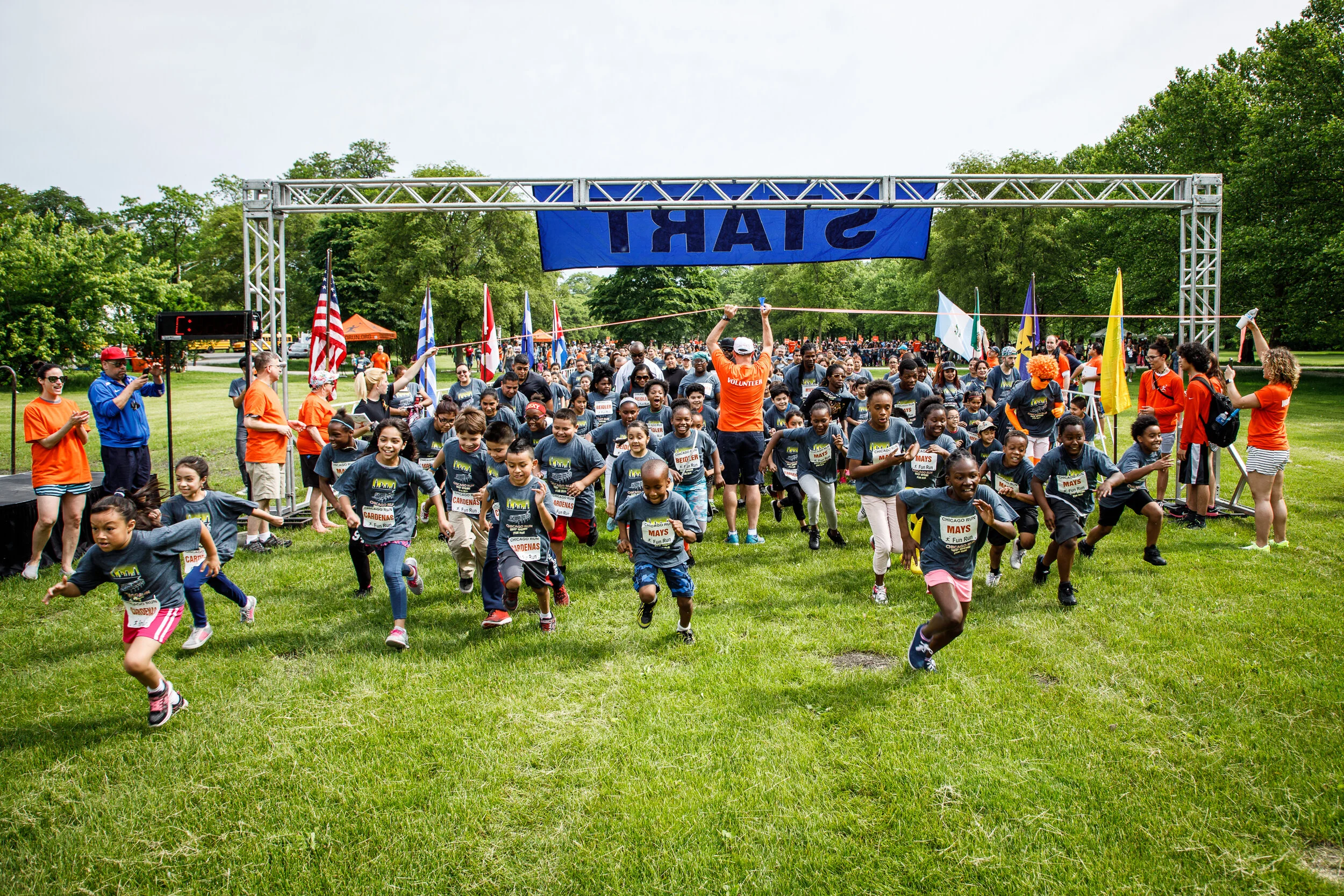 Children and adults participating in a fun run event outdoors, with participants at the starting line ready to race, some running and others cheering, with flags and a large blue banner overhead.