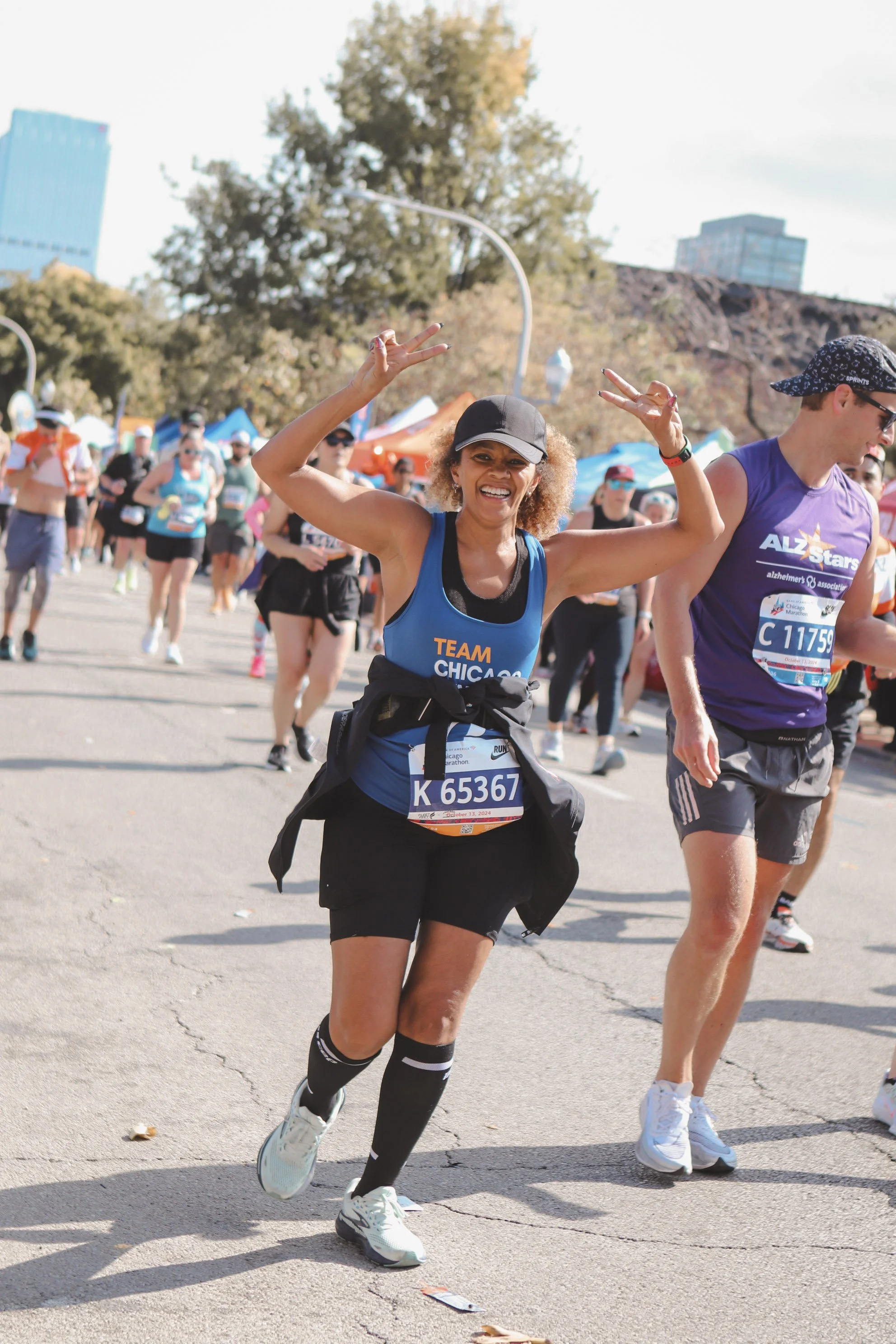 A woman smiling and making peace signs with both hands, participating in a marathon among a crowd of runners on a sunny day.