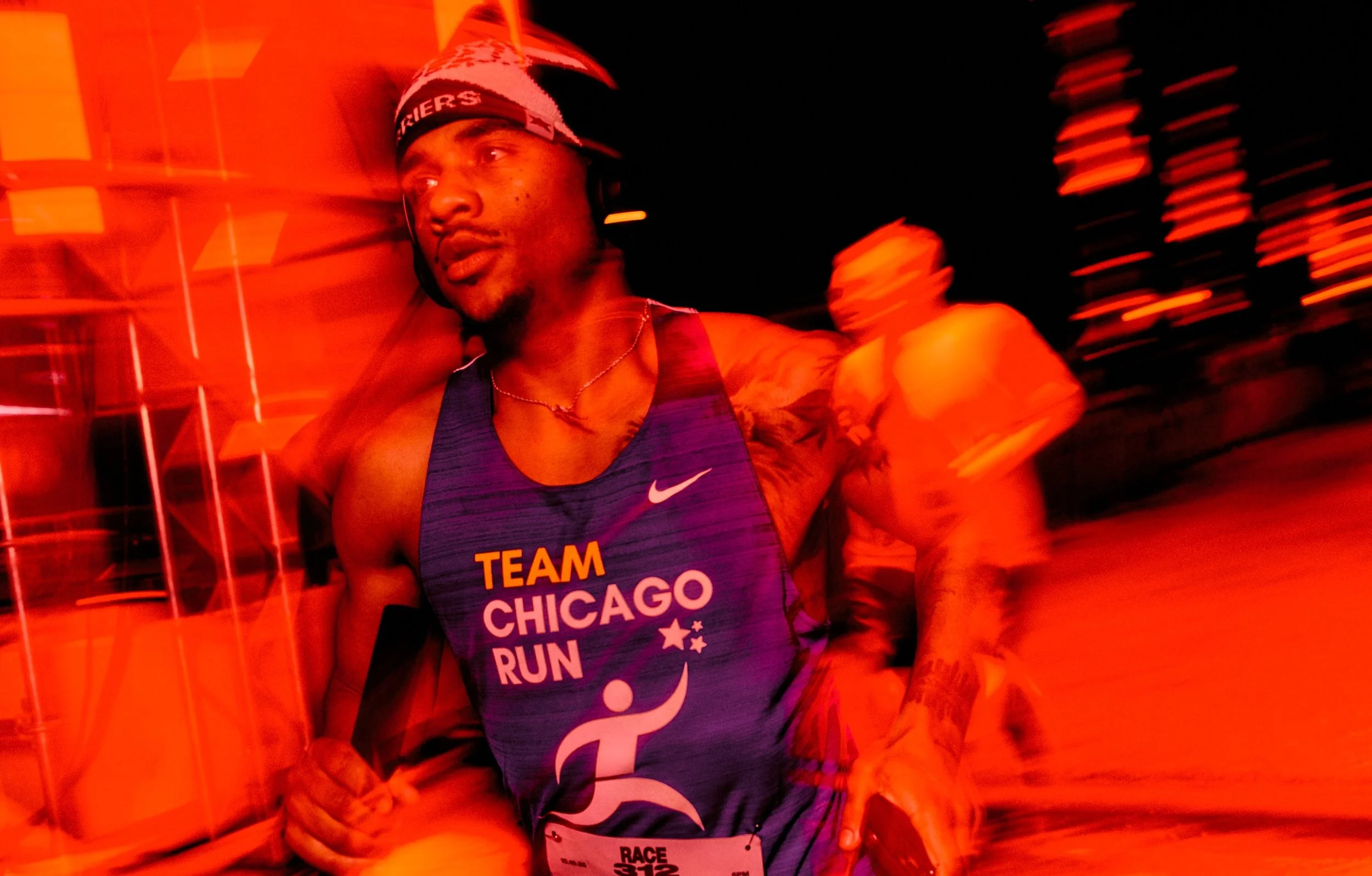 A male runner wearing a purple Nike tank top with 'Team Chicago Run' printed on it, a Santa hat, and headphones, participating in a nighttime race with blurred lights in the background.