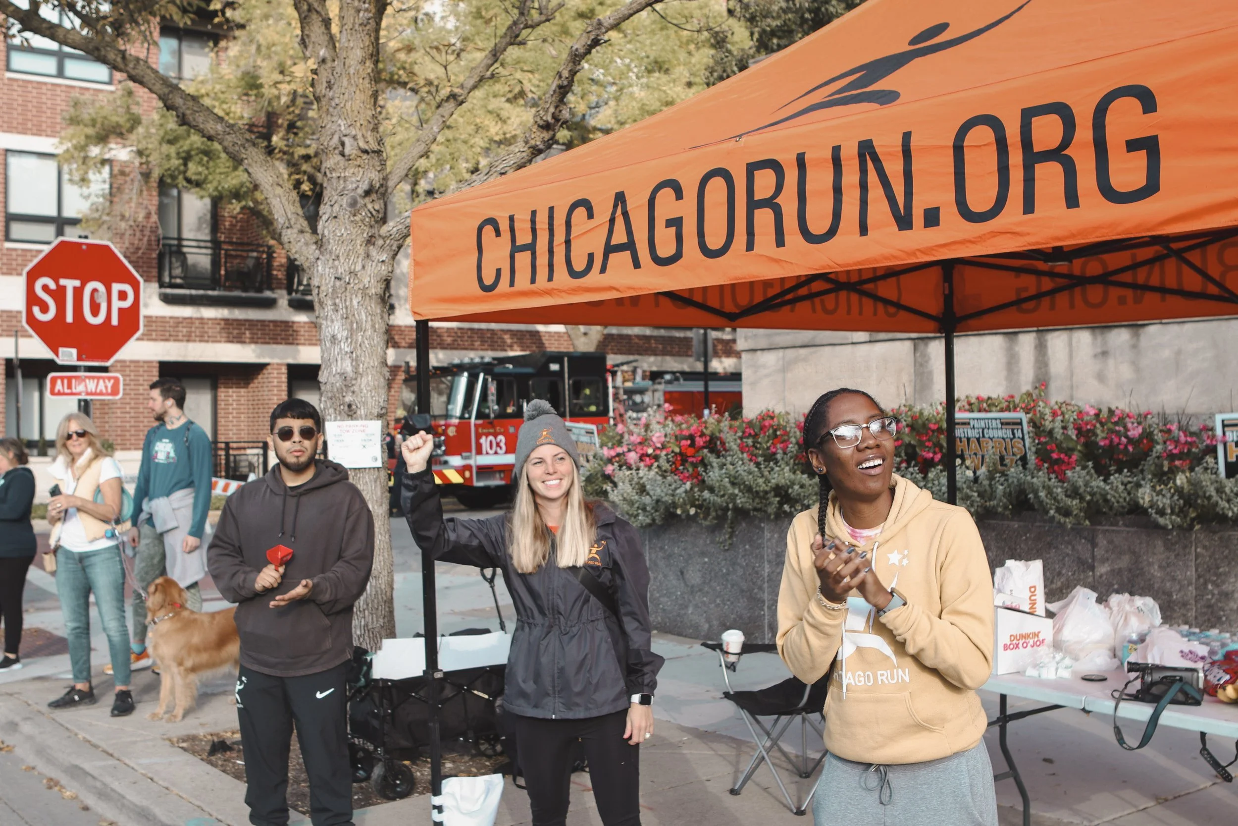 People volunteering at a Chicago Run event booth under an orange canopy with 'chicagorun.org' printed on it, on a city sidewalk with a stop sign and a fire truck in the background.