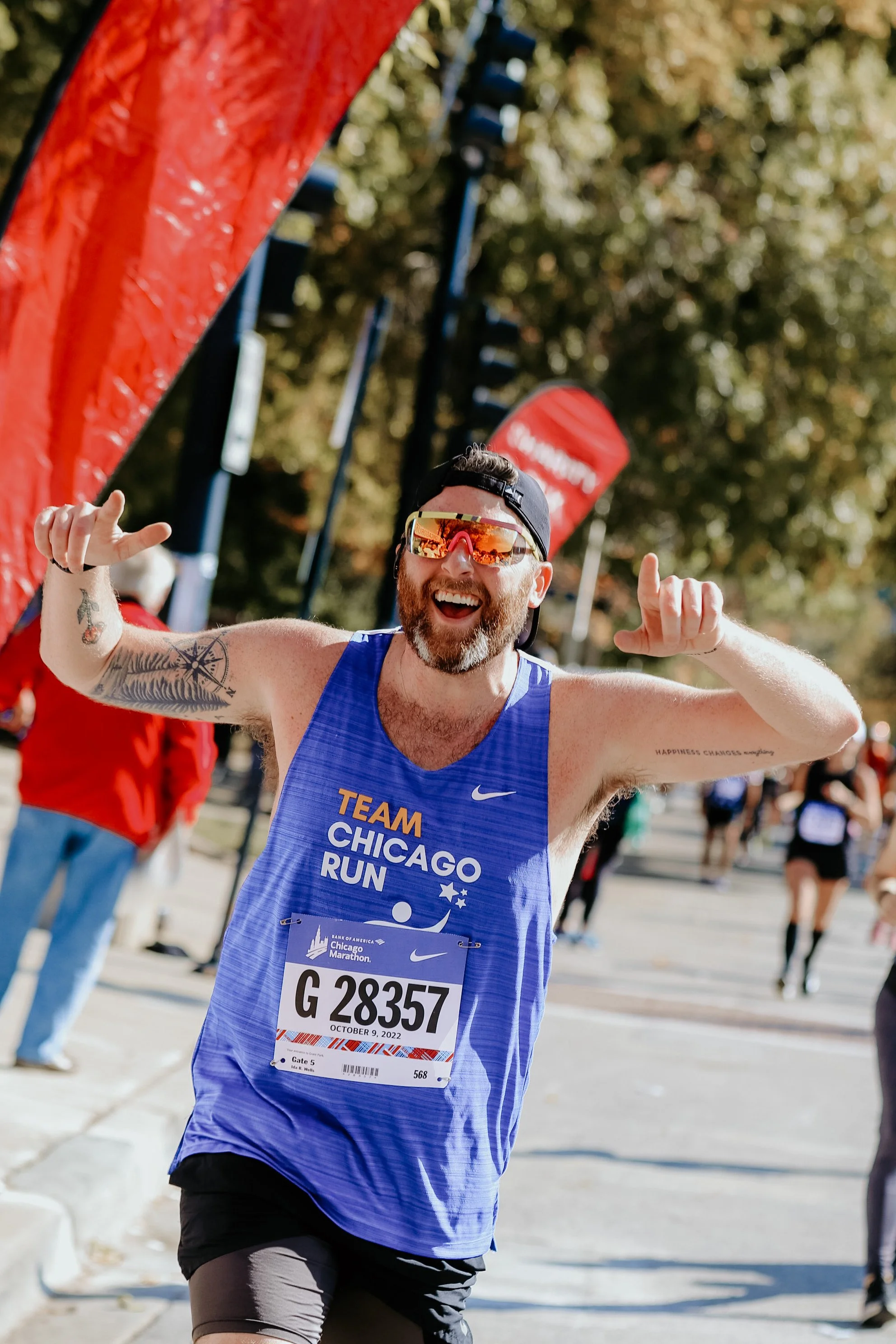 A male marathon runner wearing a blue tank top, black shorts, and sunglasses, smiling and celebrating as he crosses the finish line of the Chicago Marathon.