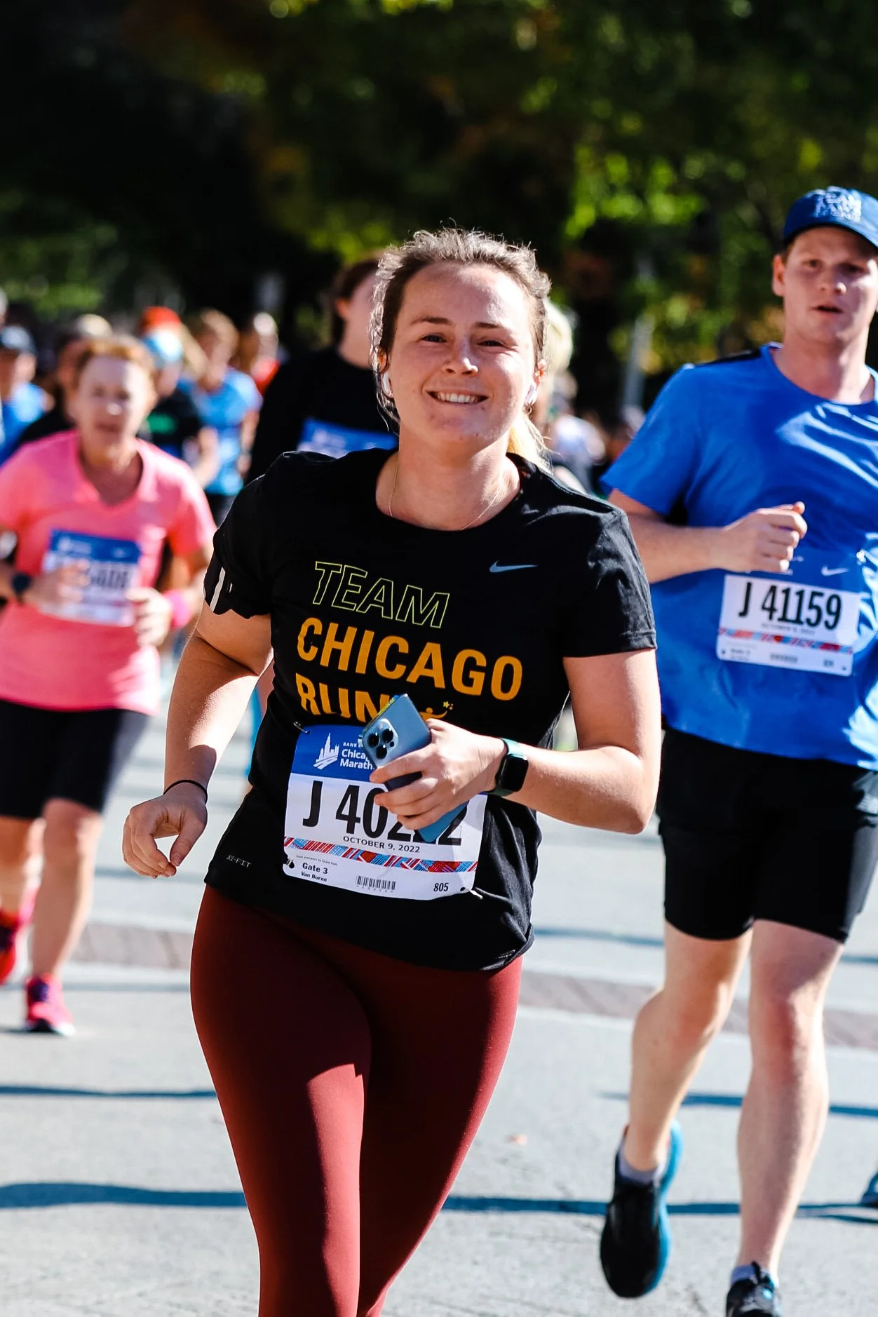 A woman running in a marathon, smiling, wearing a black shirt with yellow and orange text that says 'TEAM CHICAGO RUN'. She is holding a phone and wearing a race bib with the number J 40262. There are other runners around her, and trees in the background.
