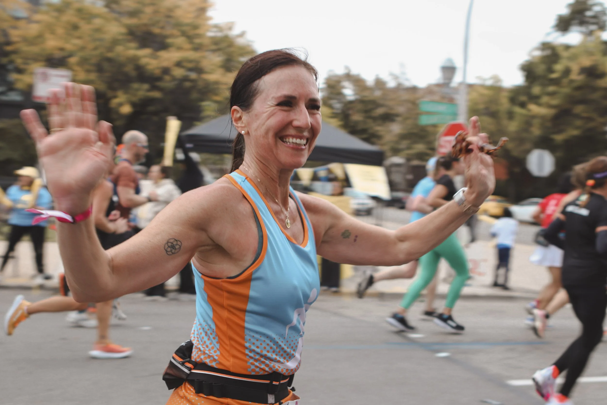 A smiling woman running in a marathon, wearing a blue and orange athletic tank top, with arms raised in a celebratory gesture, surrounded by other runners on a city street during daytime.