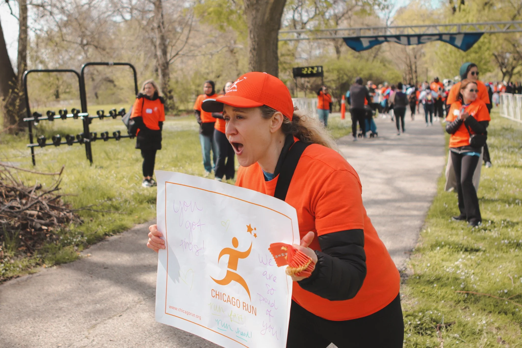 A woman in an orange shirt and cap at a charity run event, holding a sign with handwritten messages and the logo for Chicago Run, with other participants and a finish line in the background.