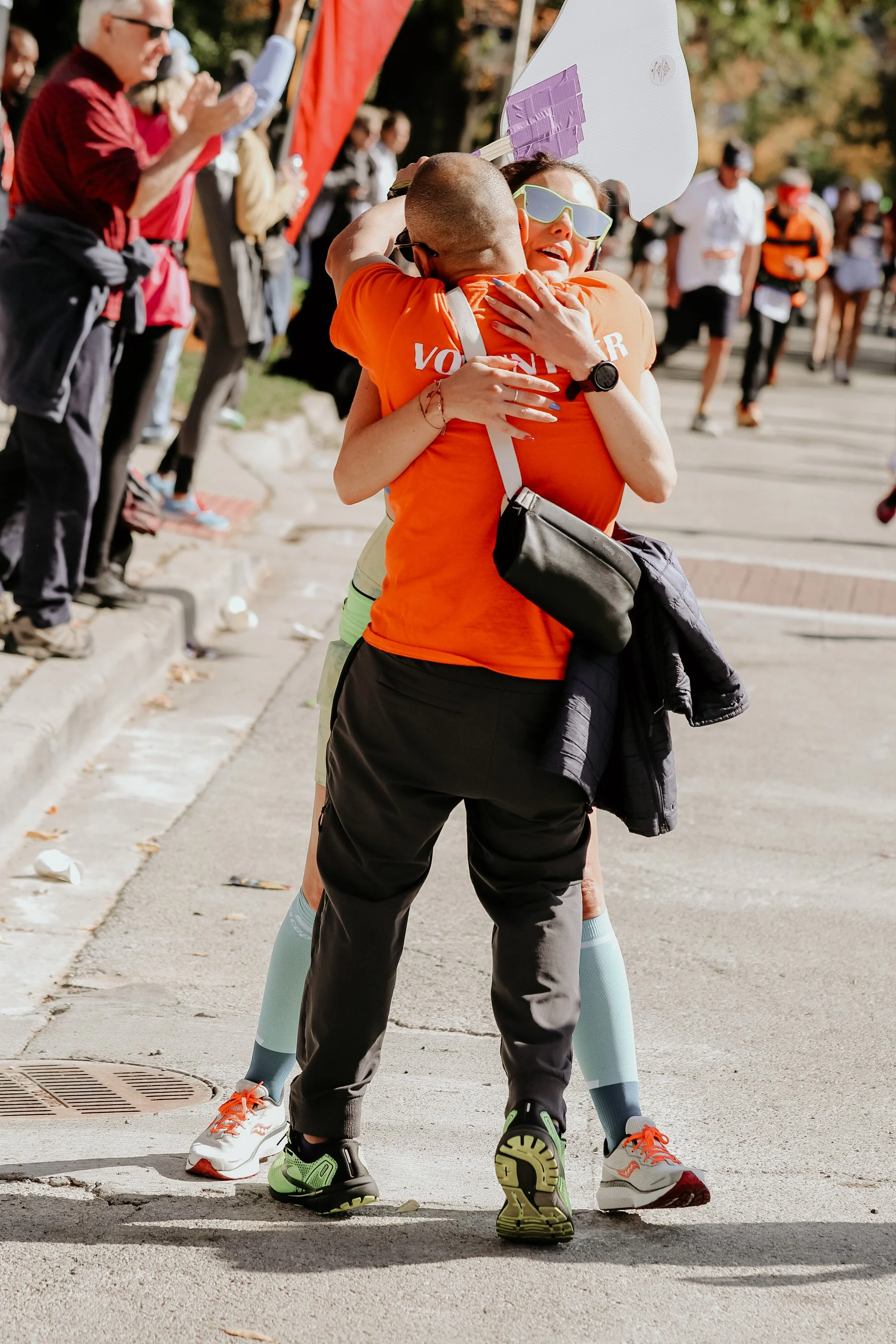 A woman with sunglasses embraces a man during a race event, with spectators cheering in the background.