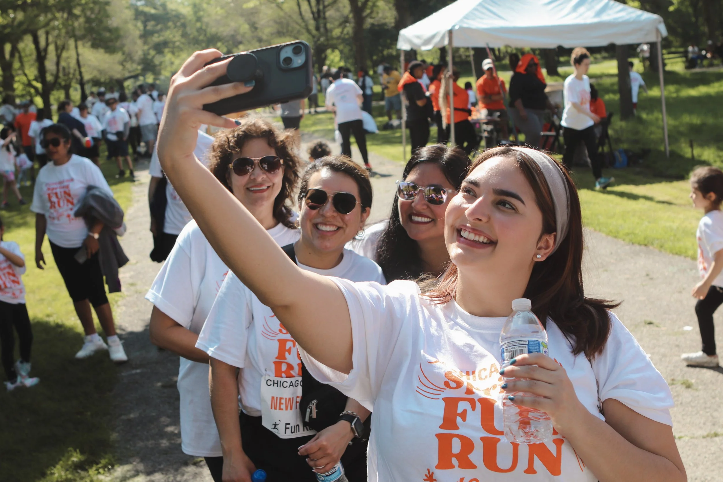 A group of women taking a selfie at a fun run event outdoors, with a crowd of other participants and a white tent in the background.