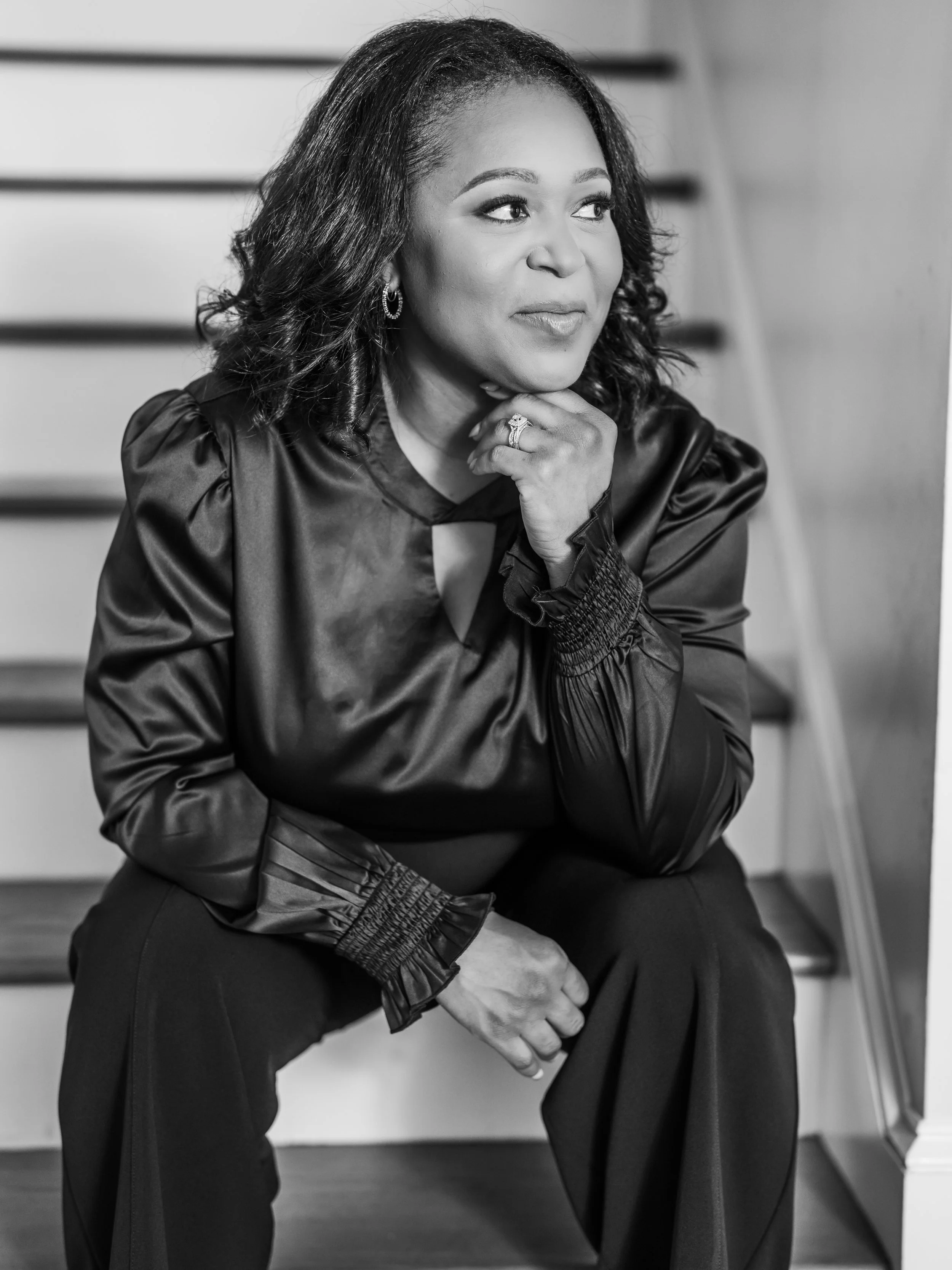 A portrait of a woman sitting on stairs, with her chin resting on her hand, looking to the side, dressed in a silky blouse and pants, in black and white.
