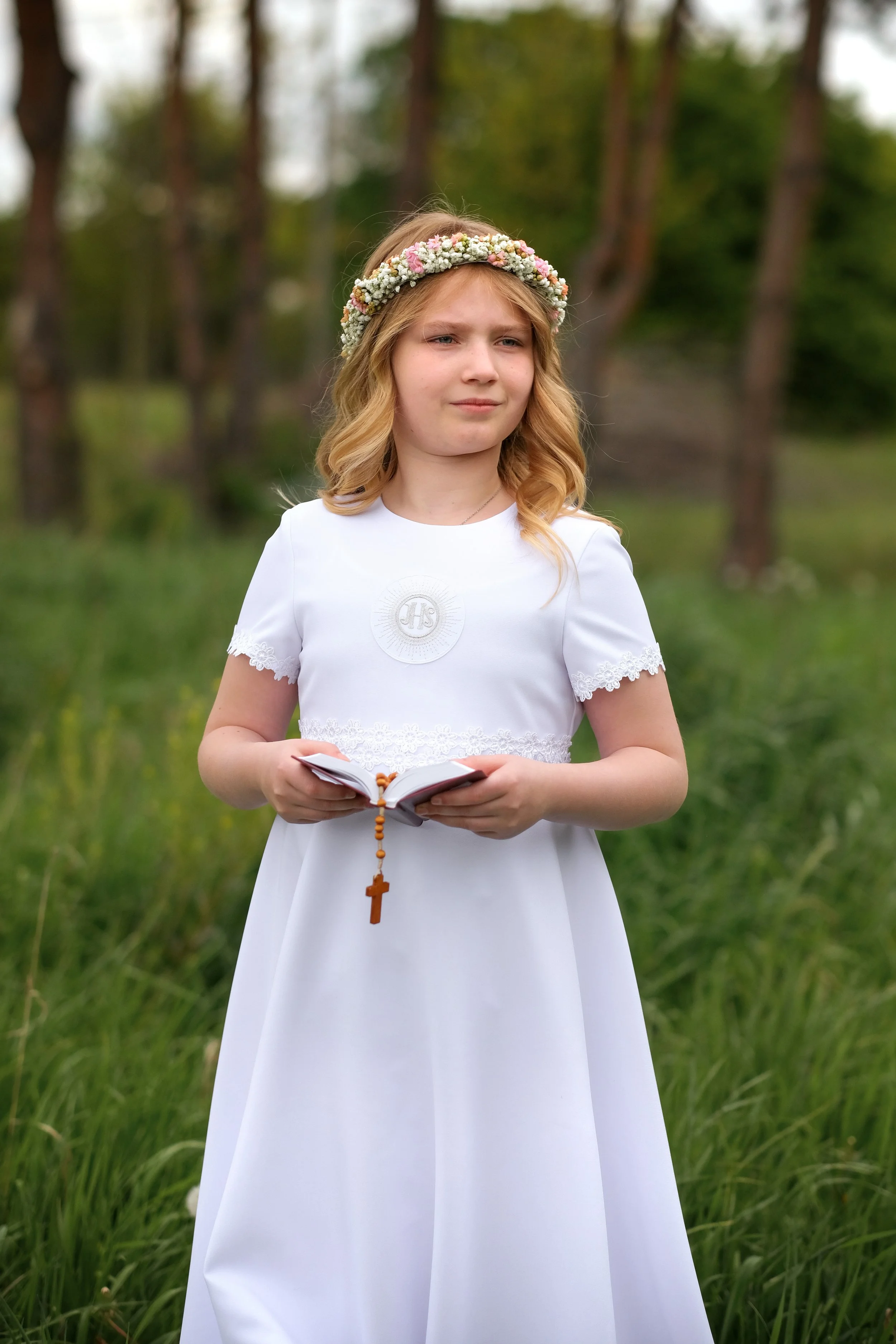 A young girl wearing a white dress and a flower crown holding a prayer book with a rosary in an outdoor setting with green grass and trees in the background.