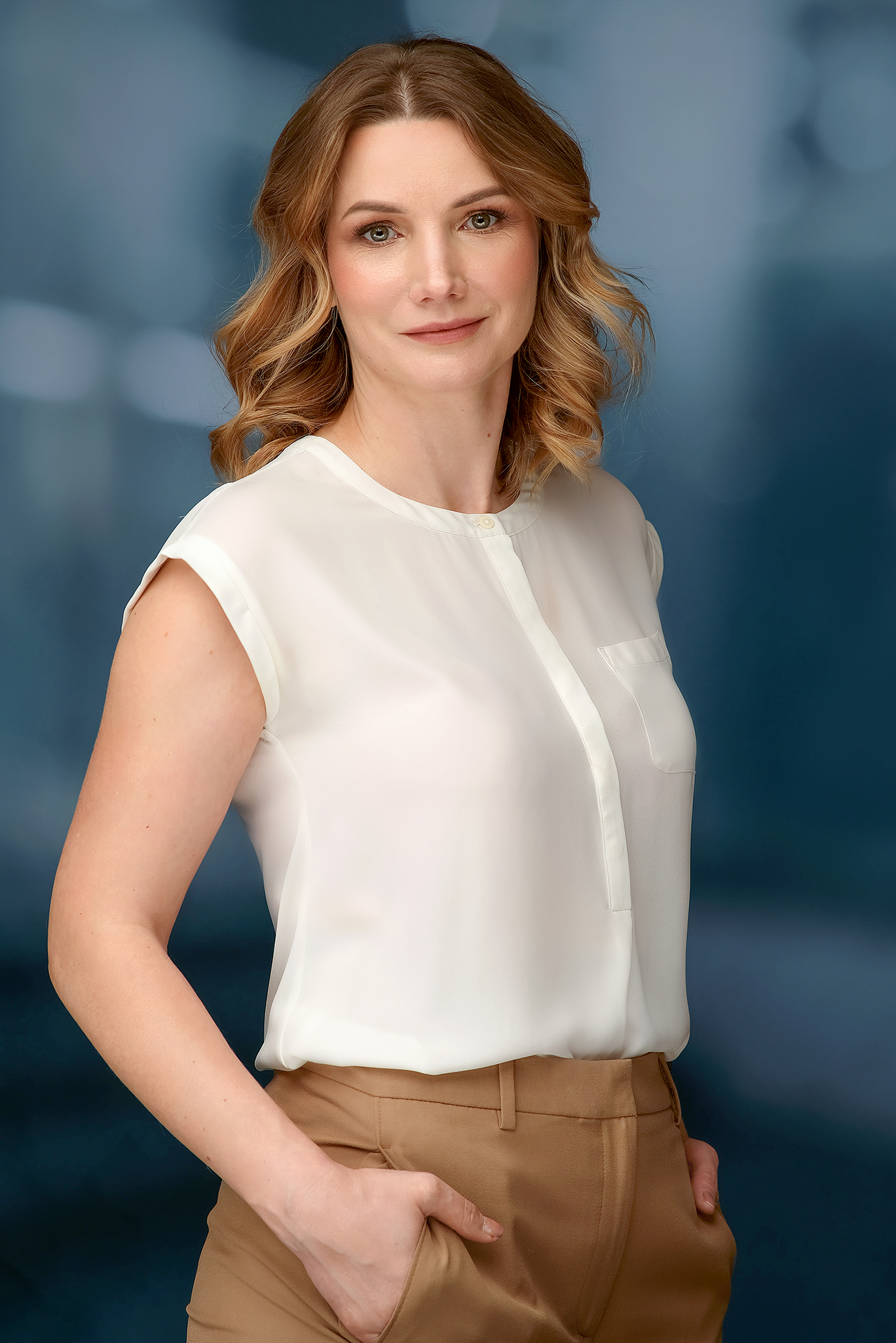 A woman with shoulder-length wavy brown hair, wearing a white sleeveless blouse and tan pants, posing against a blurred blue background.