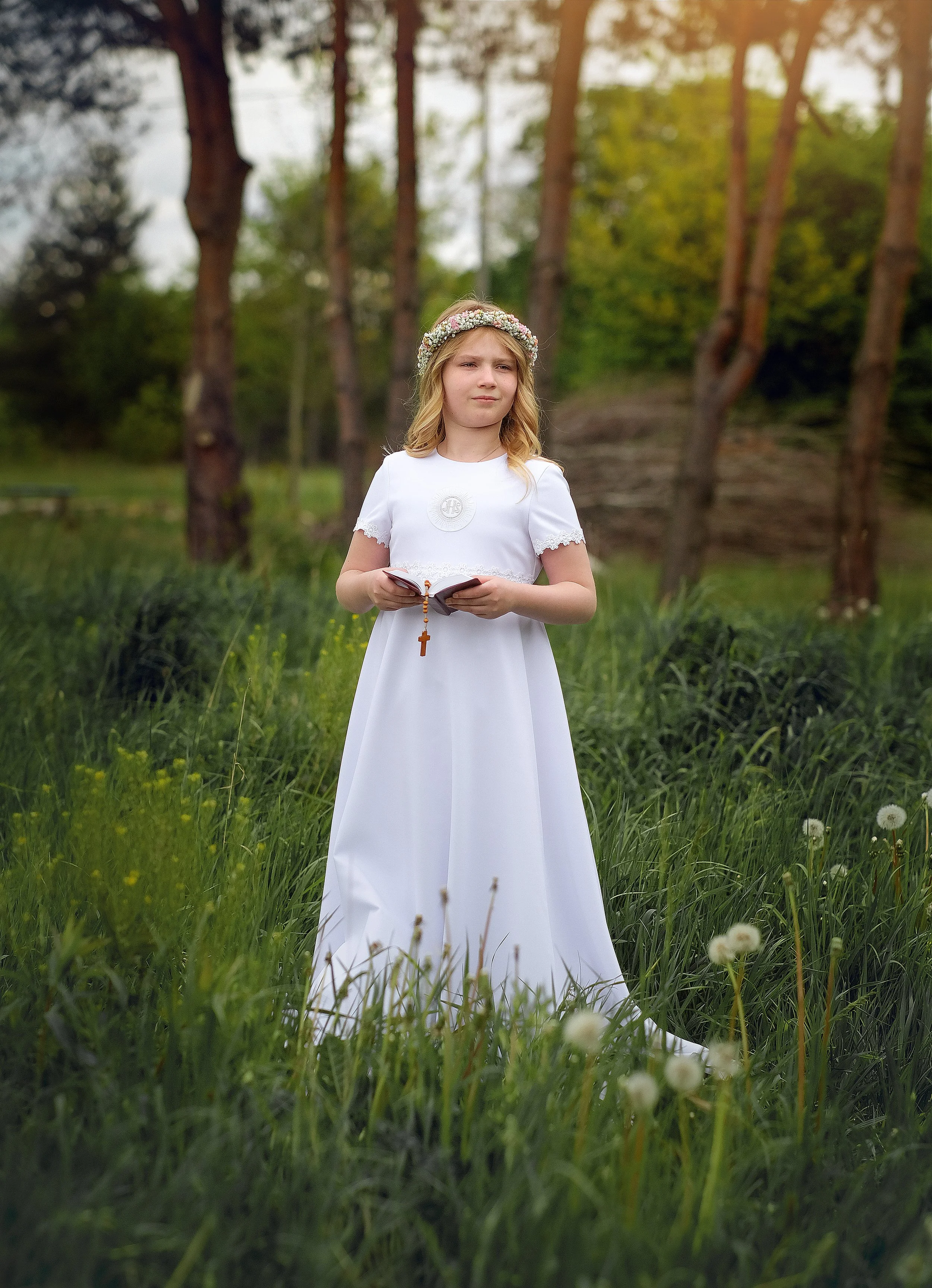 A young girl dressed in a white dress with a floral crown, holding a rosary, standing in a grassy field surrounded by trees.