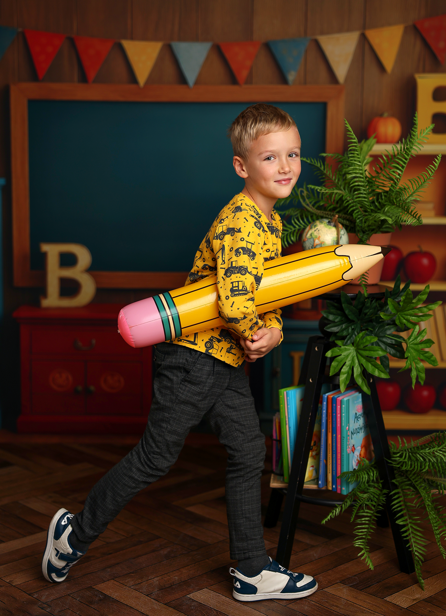 A young boy in a yellow shirt with construction vehicle patterns, holding a large yellow and pink crayon, standing in a room decorated with colorful triangle banners, greenery, books, pumpkins, and a chalkboard.