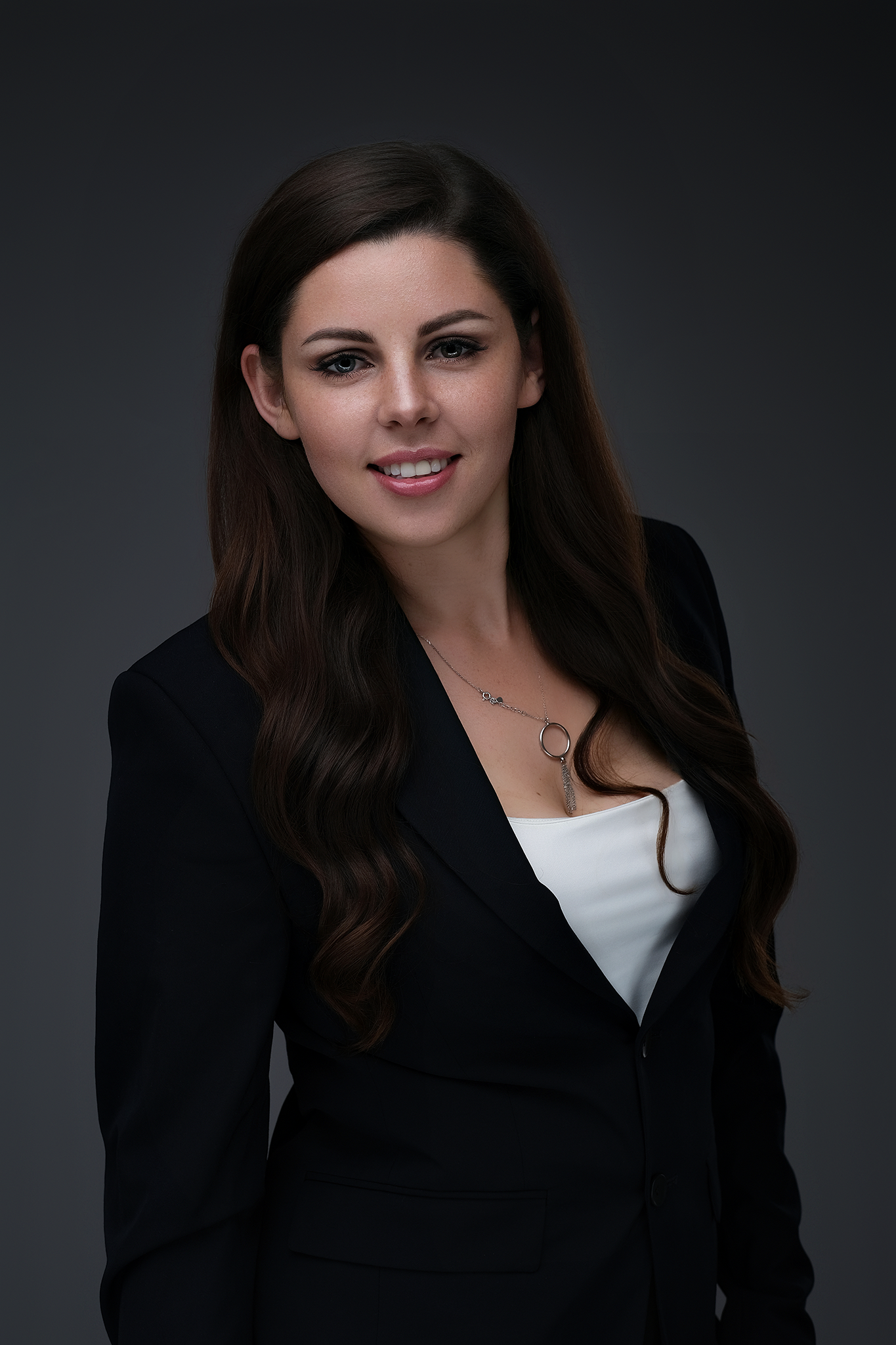 Professional woman with long dark hair, wearing a black blazer and white top, posing against a dark gray gradient background.