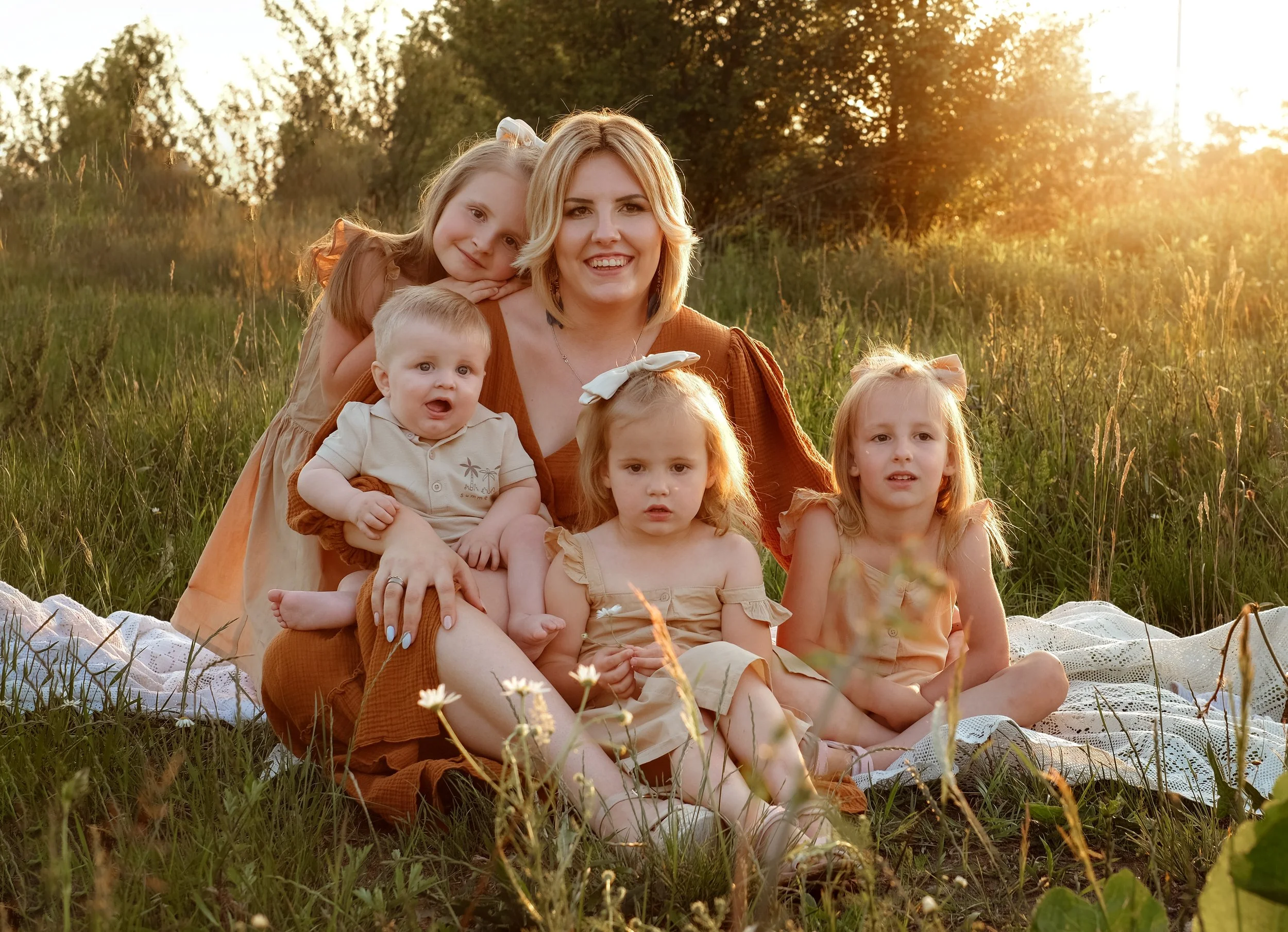 A woman and five young girls sitting on a blanket in a field during sunset, smiling and posing for the photo.