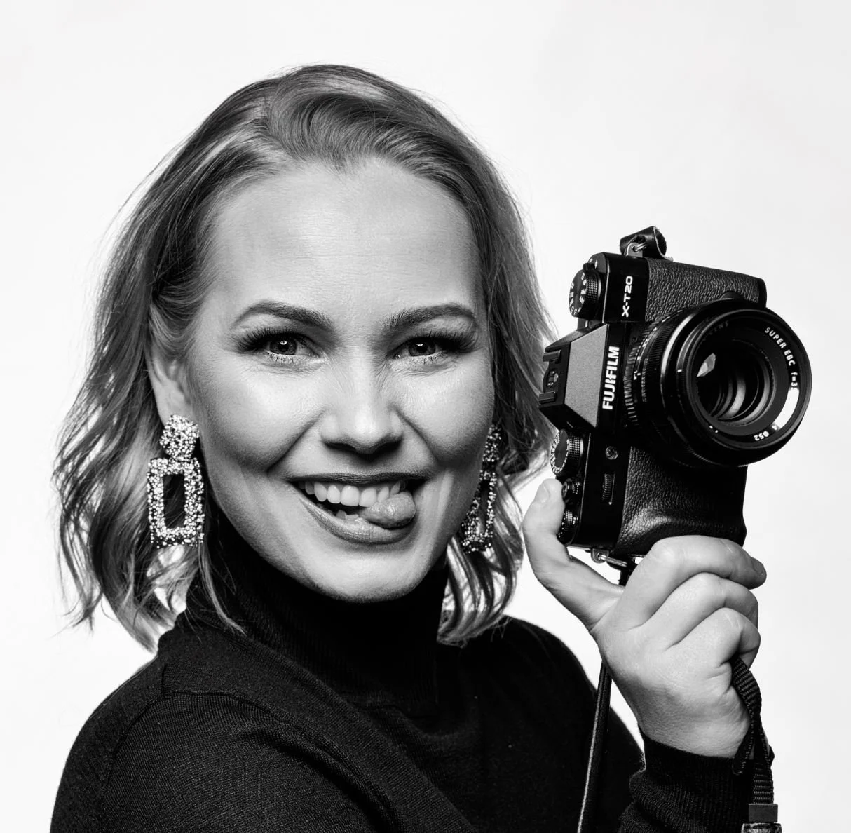 Black and white portrait of a smiling woman with wavy blonde hair, wearing large earrings, holding a Fujifilm camera near her face, with her tongue slightly out.
