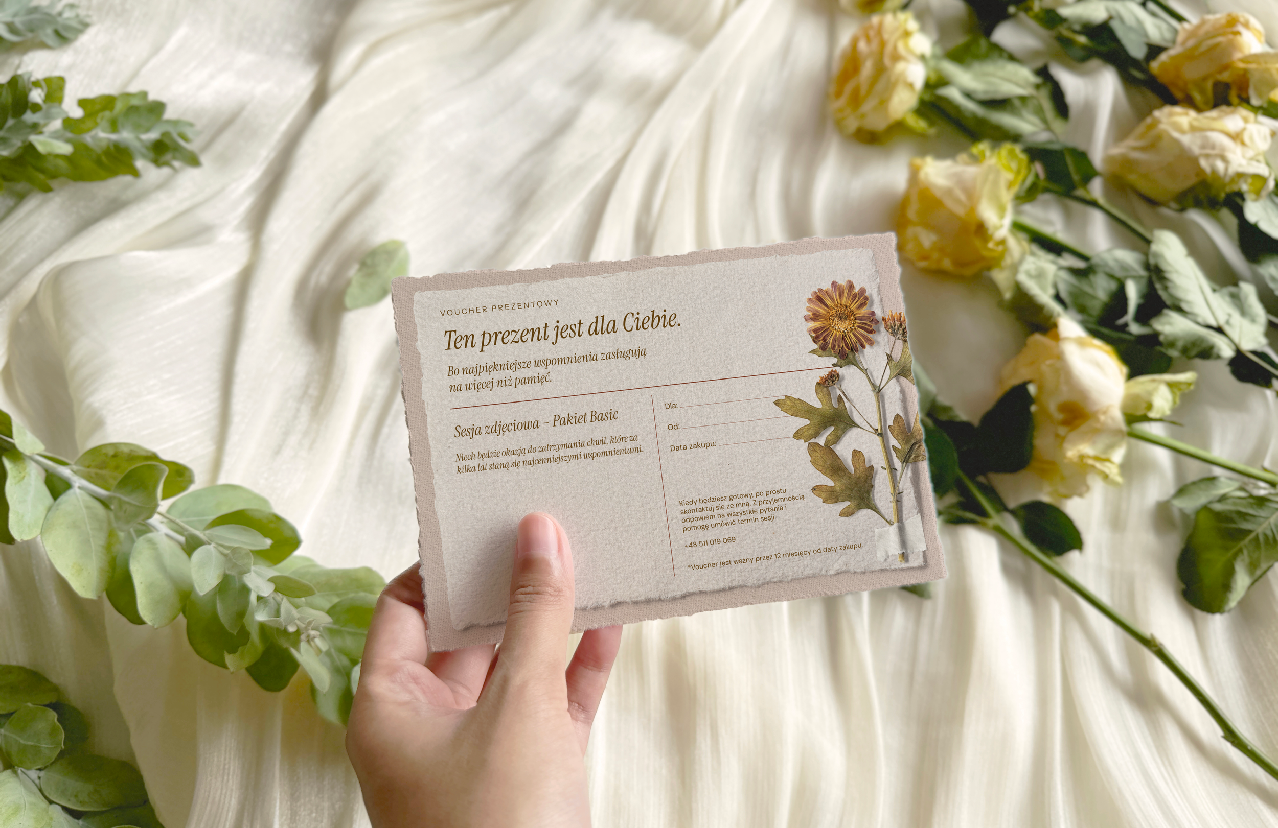 Close-up of a hand holding a gift voucher with floral decorations on a soft fabric surface, surrounded by yellow and white flowers and green leaves.