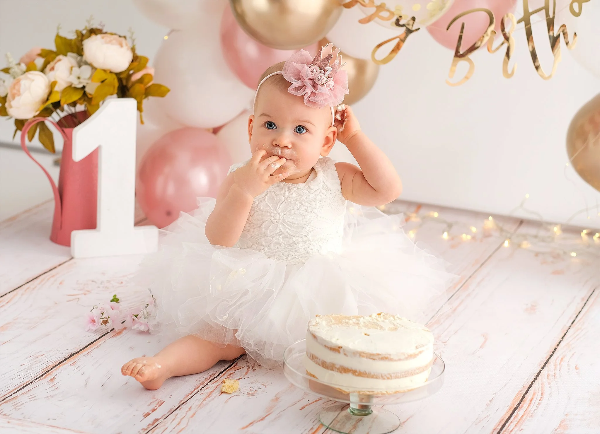 A baby girl celebrating her first birthday, sitting on a white wooden floor with a partially eaten cake in front of her. She is wearing a white lace dress with a fluffy tulle skirt and a pink headband with a flower and crown decoration. The backgroun