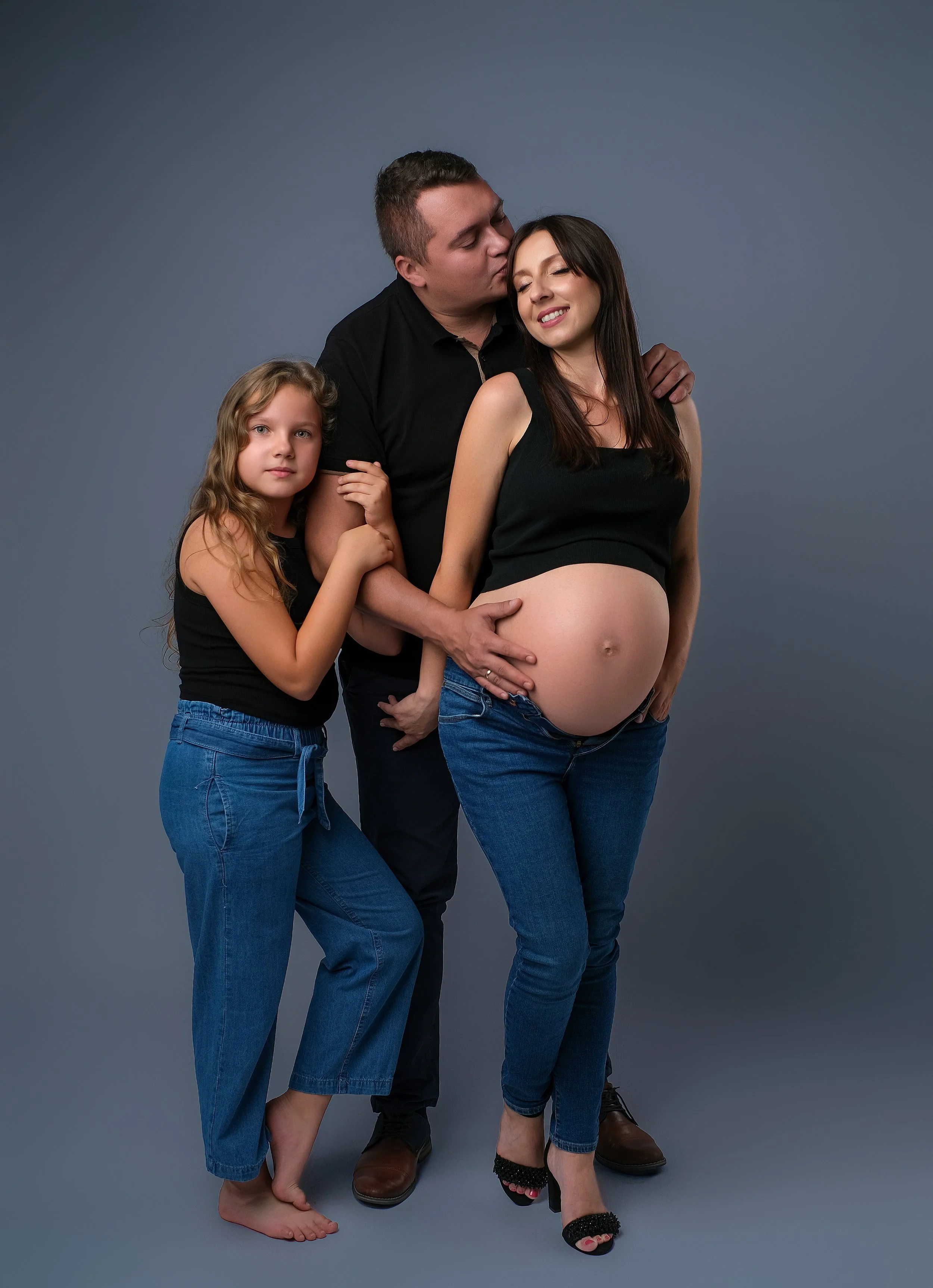 Family portrait of a pregnant woman with her partner and daughter against a gray background; the woman is smiling with her eyes closed, the man is kissing her cheek, and the daughter is holding her arm.