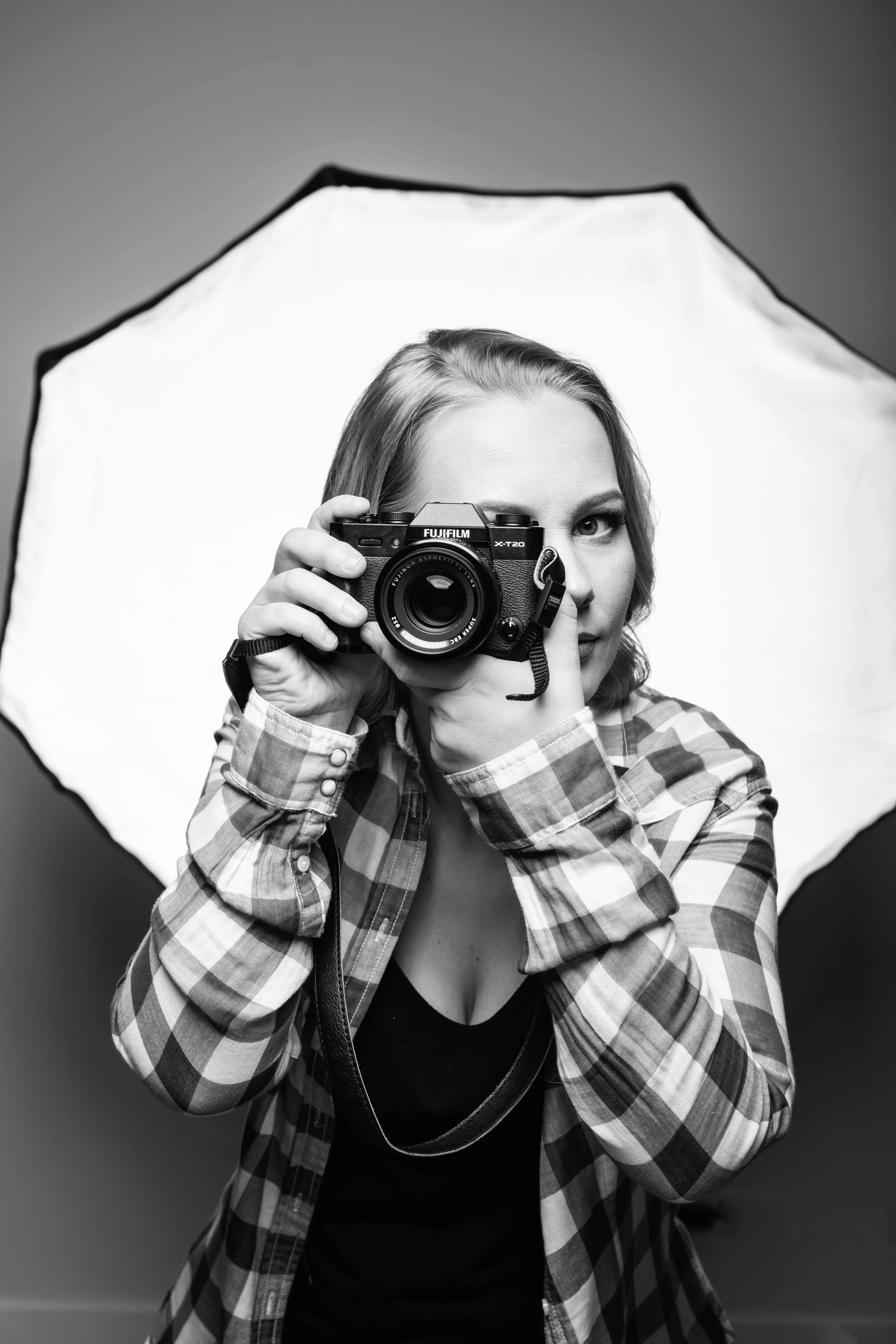 Black and white photo of a woman holding a camera up to her eye, with a photography studio light diffuser in the background.