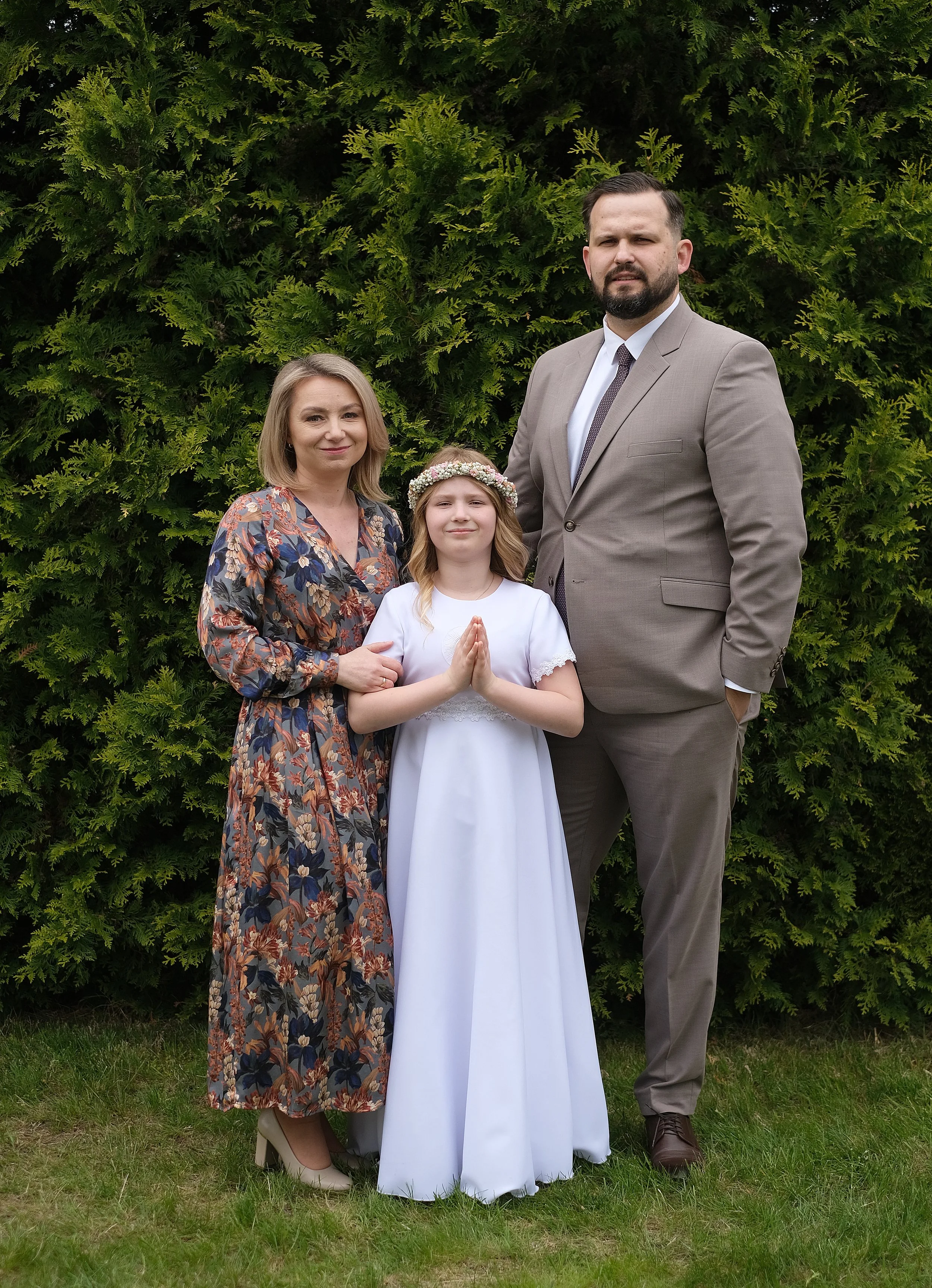 A family of three standing outdoors in front of green bushes, with a woman in a floral dress, a girl in a white dress with a floral crown, and a man in a beige suit. The girl has her hands pressed together in a prayer pose.