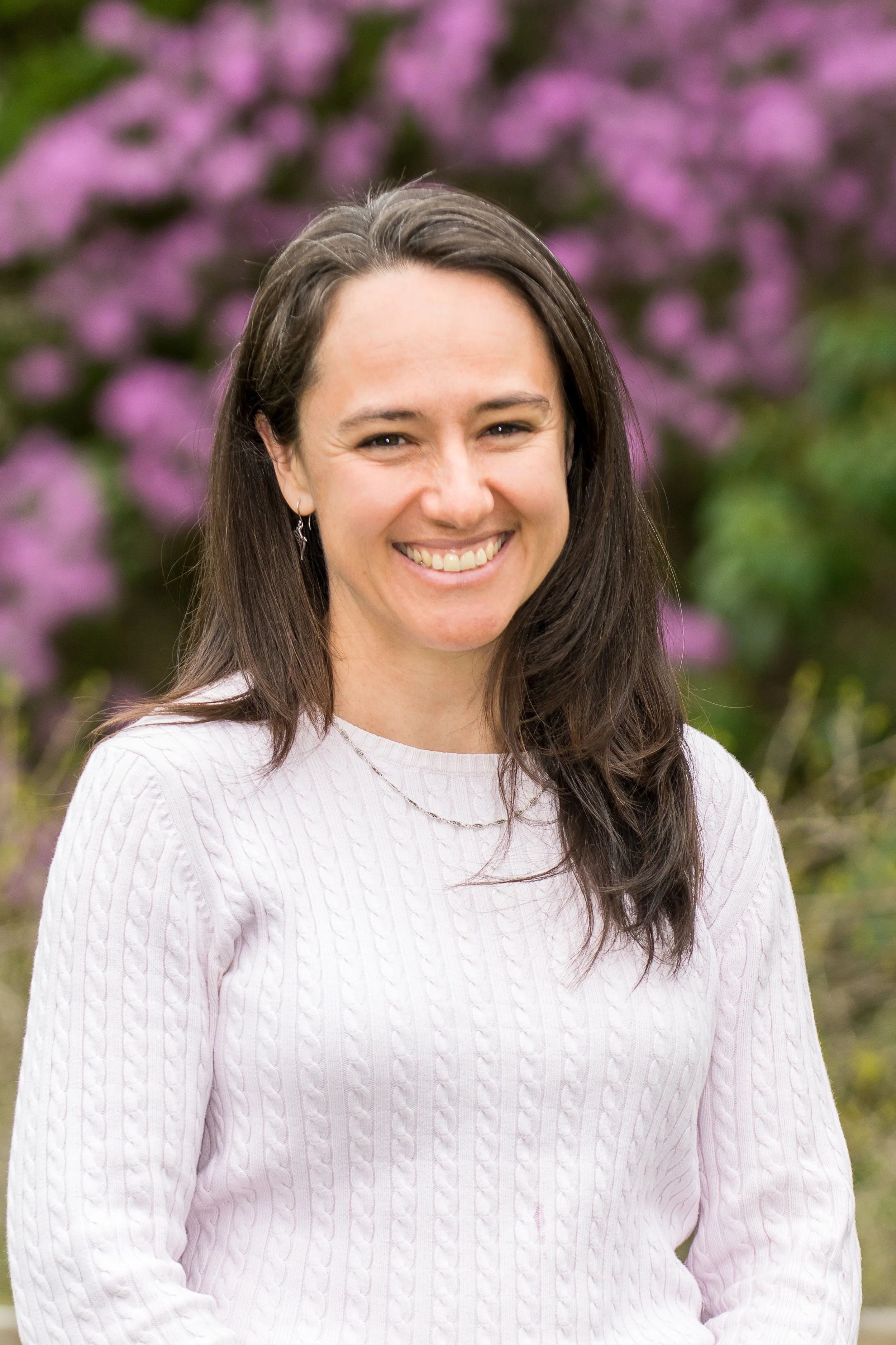 A photo of Megan in a pink sweater smiling at the camera in front of a blooming lilac bush.
