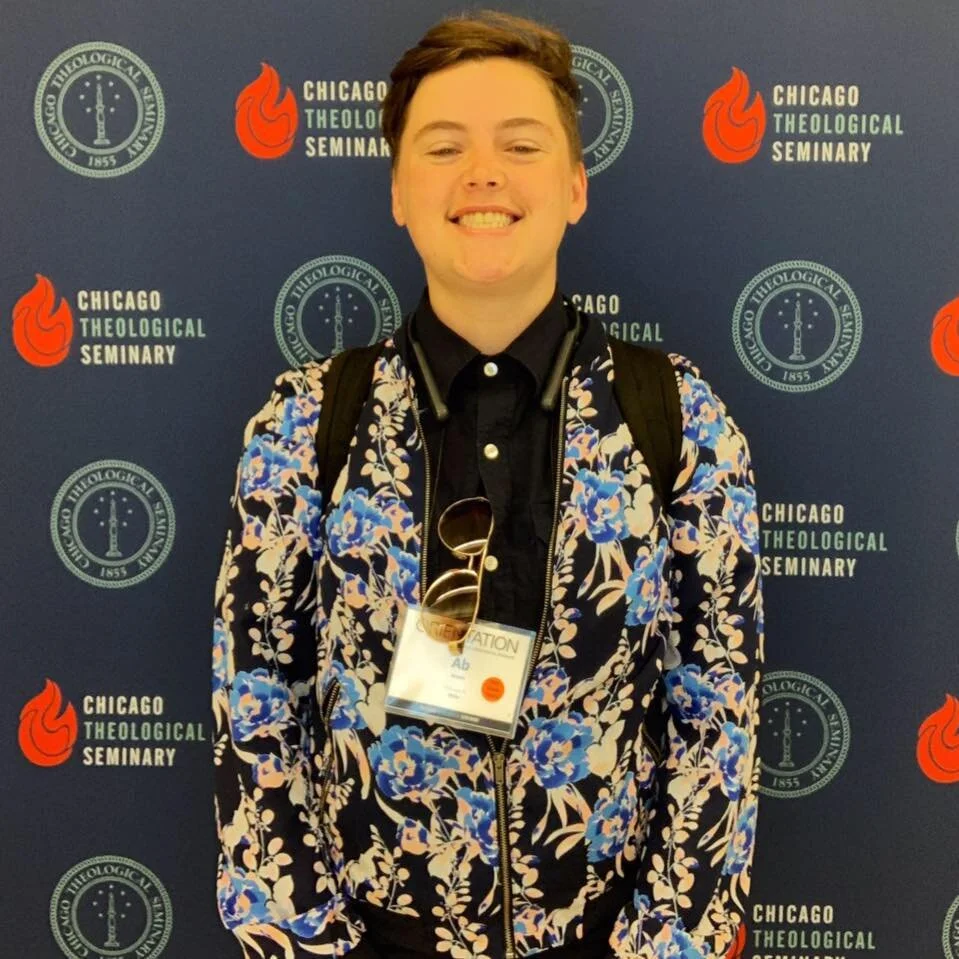 A photo of Ab in a flora jacket smiling in front of a Chicago Theological Seminary banner.