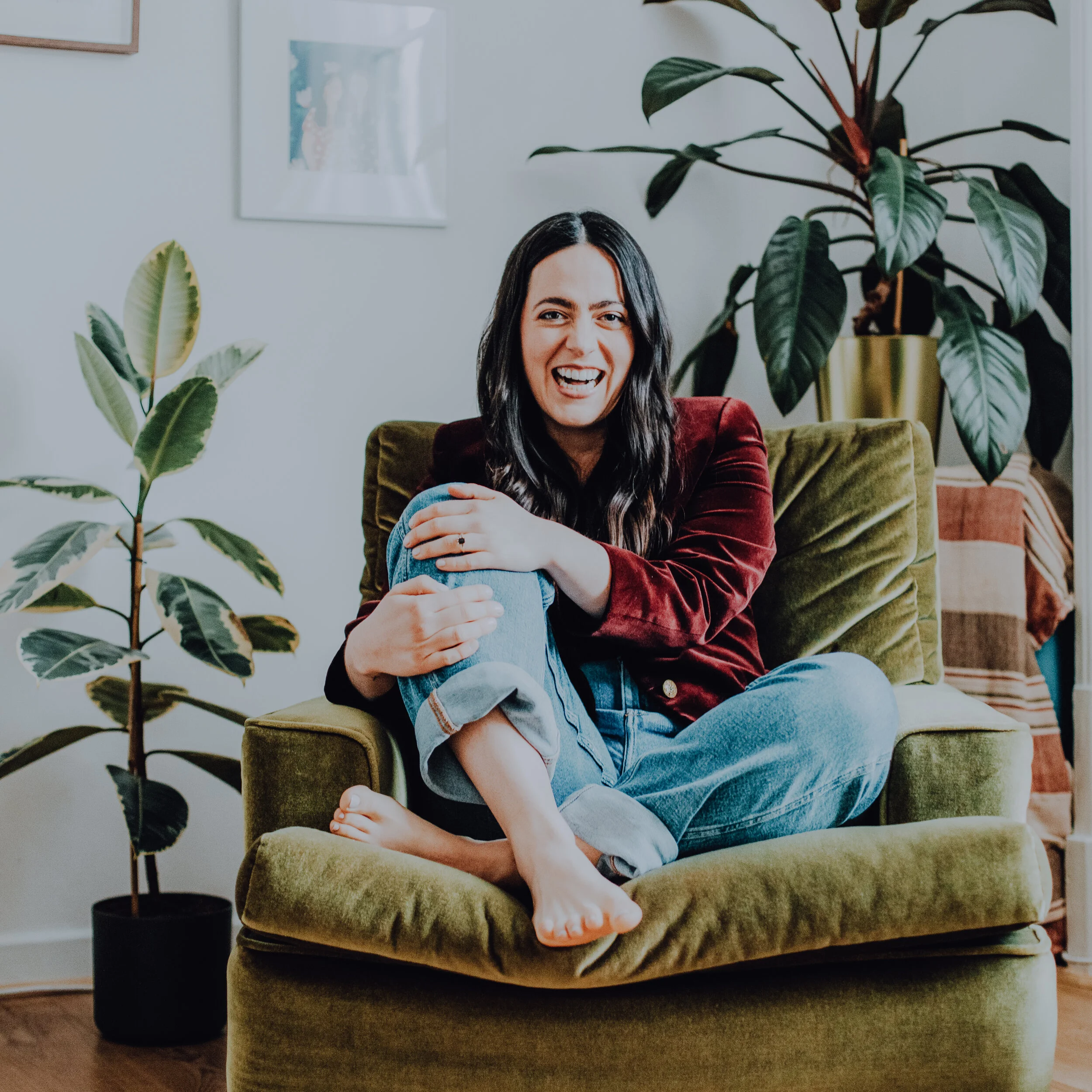 A photo of Allison sitting in a green velvet chair with a her legs crossed in front of her, surrounded by plants.