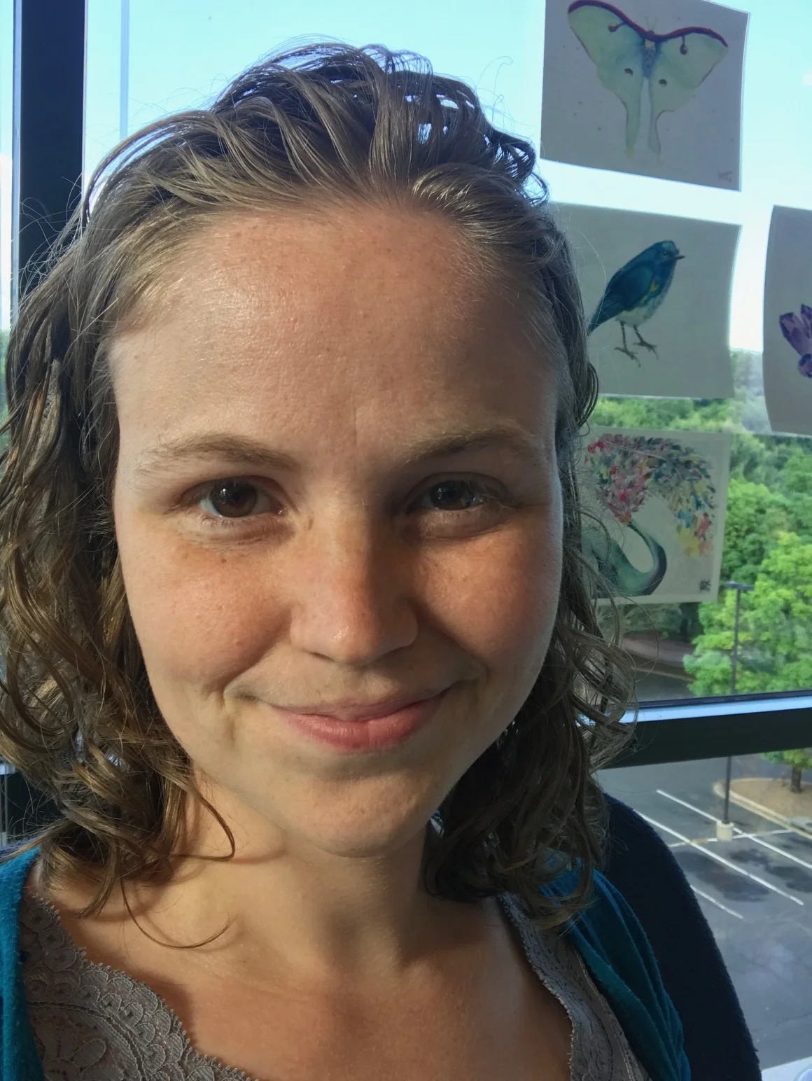 A selfie by Amanda smiling into the camera with illustrations of birds and butterflies taped to the window back and to the right.