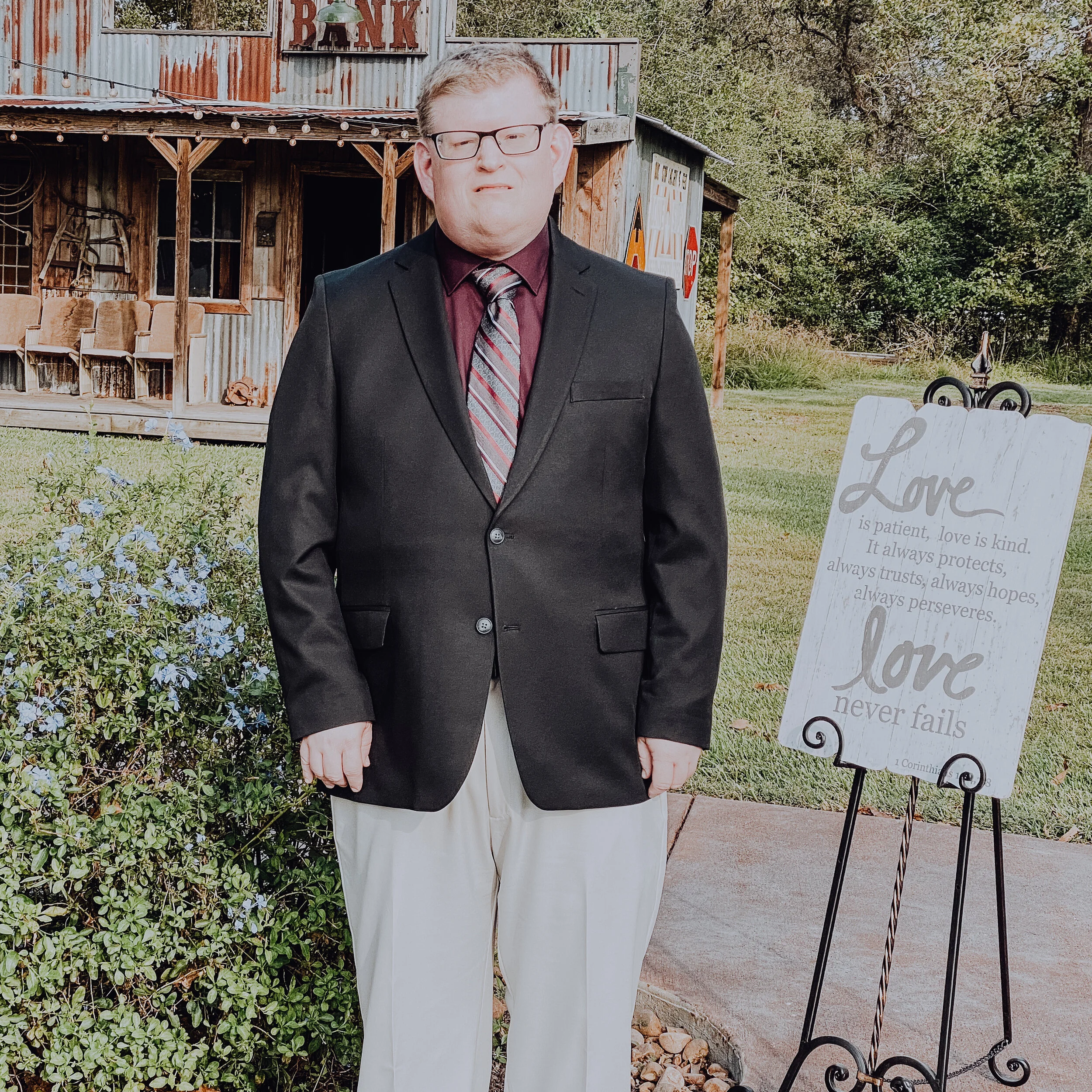 A photo of Andy in a suit next to a sign that quotes I Corinthians 13. He is standing on a sidewalk in front of a rustic-looking building with an old "bank sign" on its parapet.